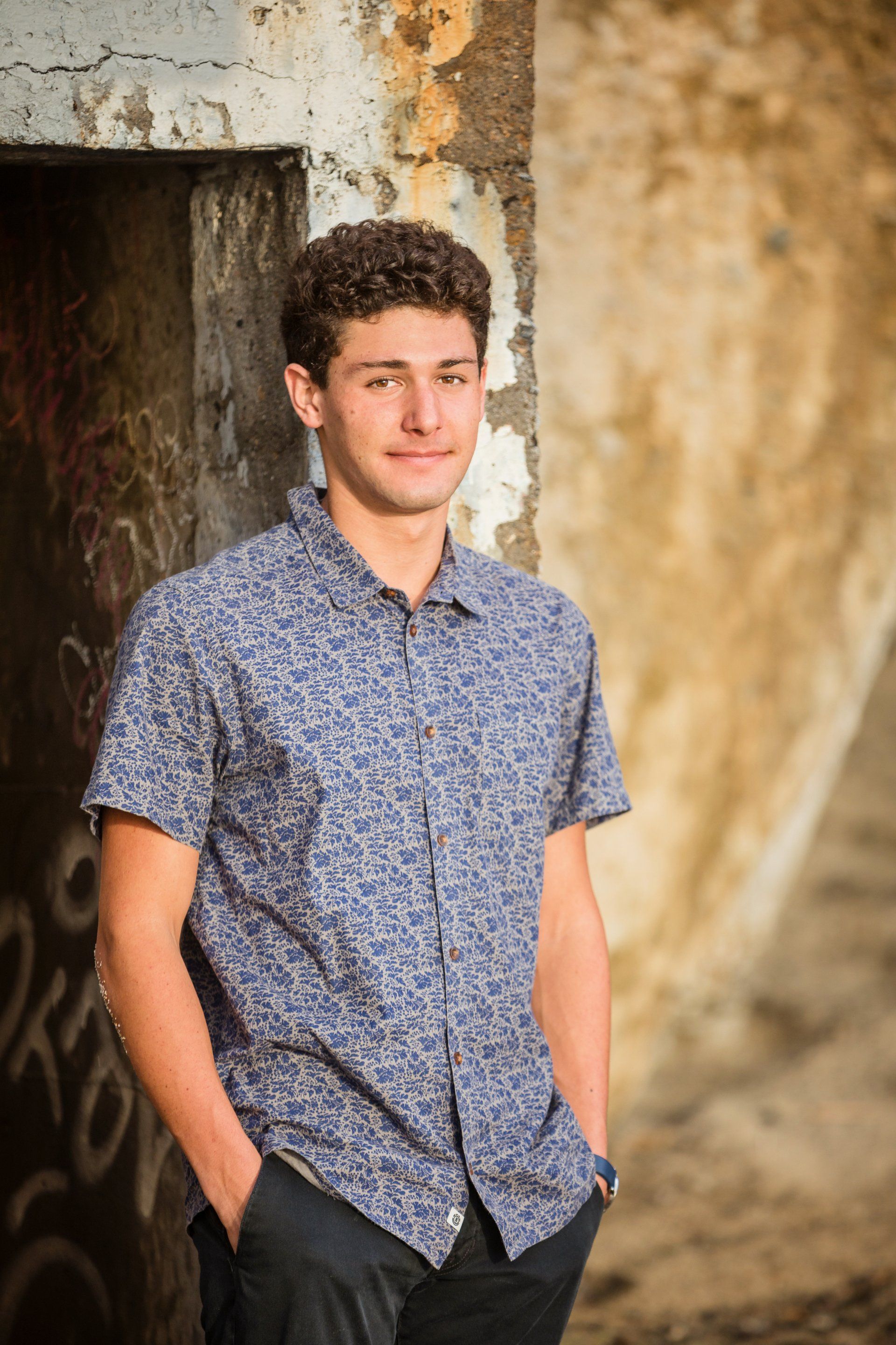 A young man in a blue shirt is standing in front of a wall with his hands in his pockets.