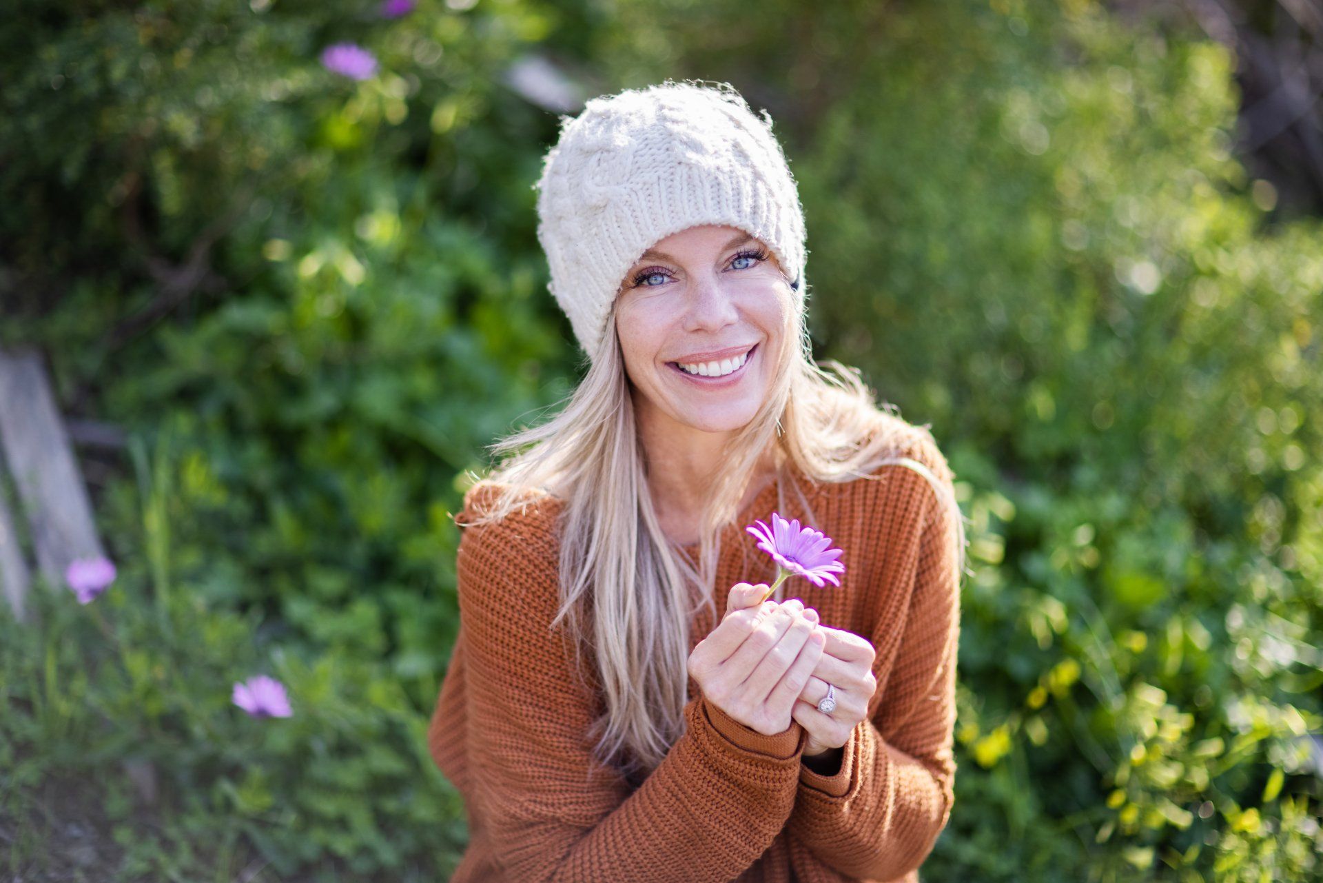 A woman wearing a beanie and a sweater is holding a flower in her hands.