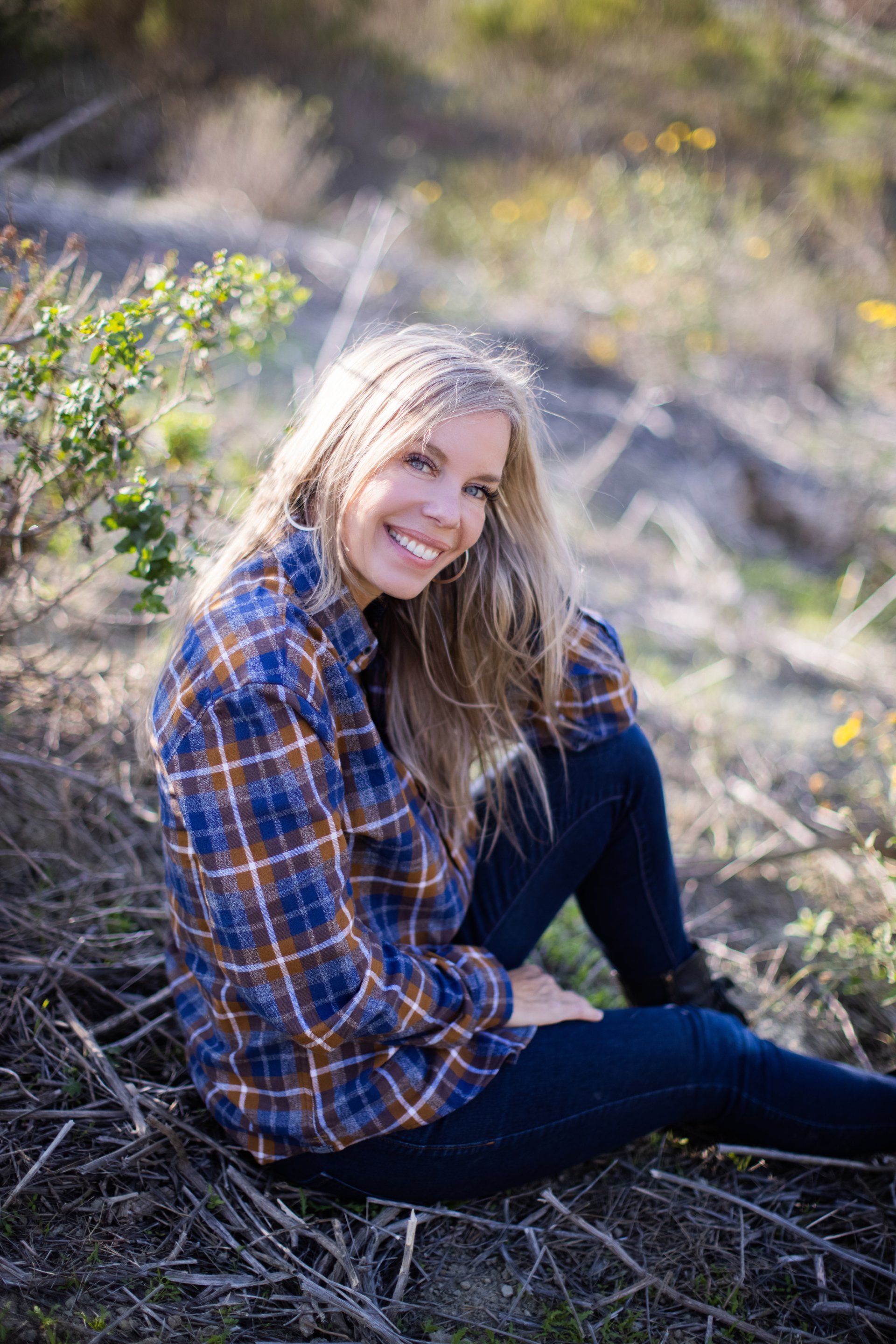 A woman is sitting on the ground wearing a plaid shirt and smiling.