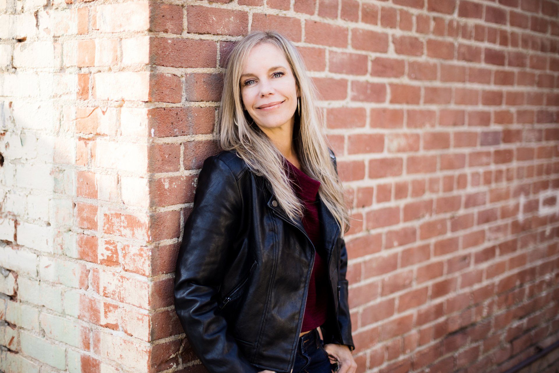 A woman in a leather jacket is leaning against a brick wall.
