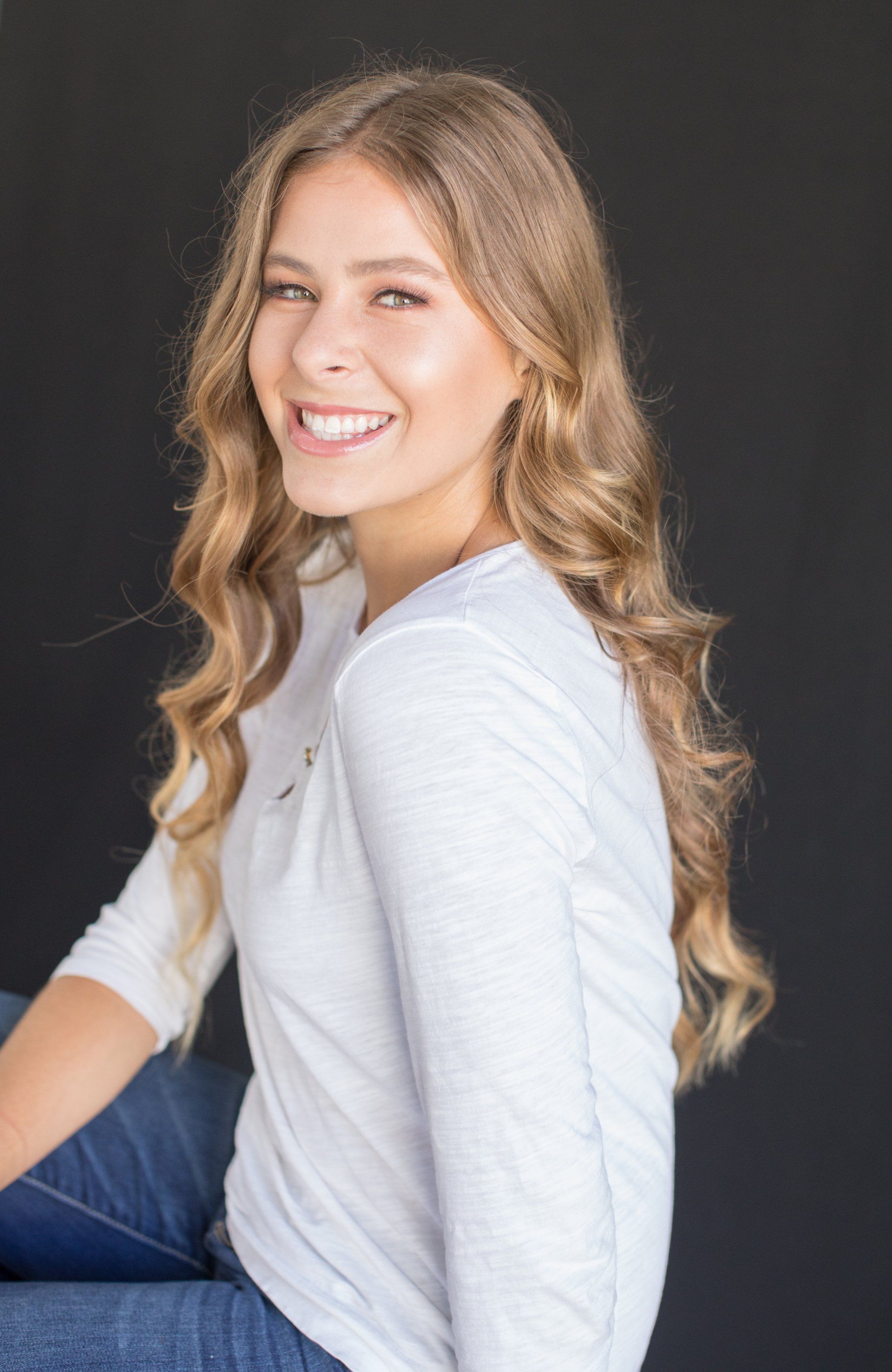A young woman with long blonde hair is sitting down and smiling.