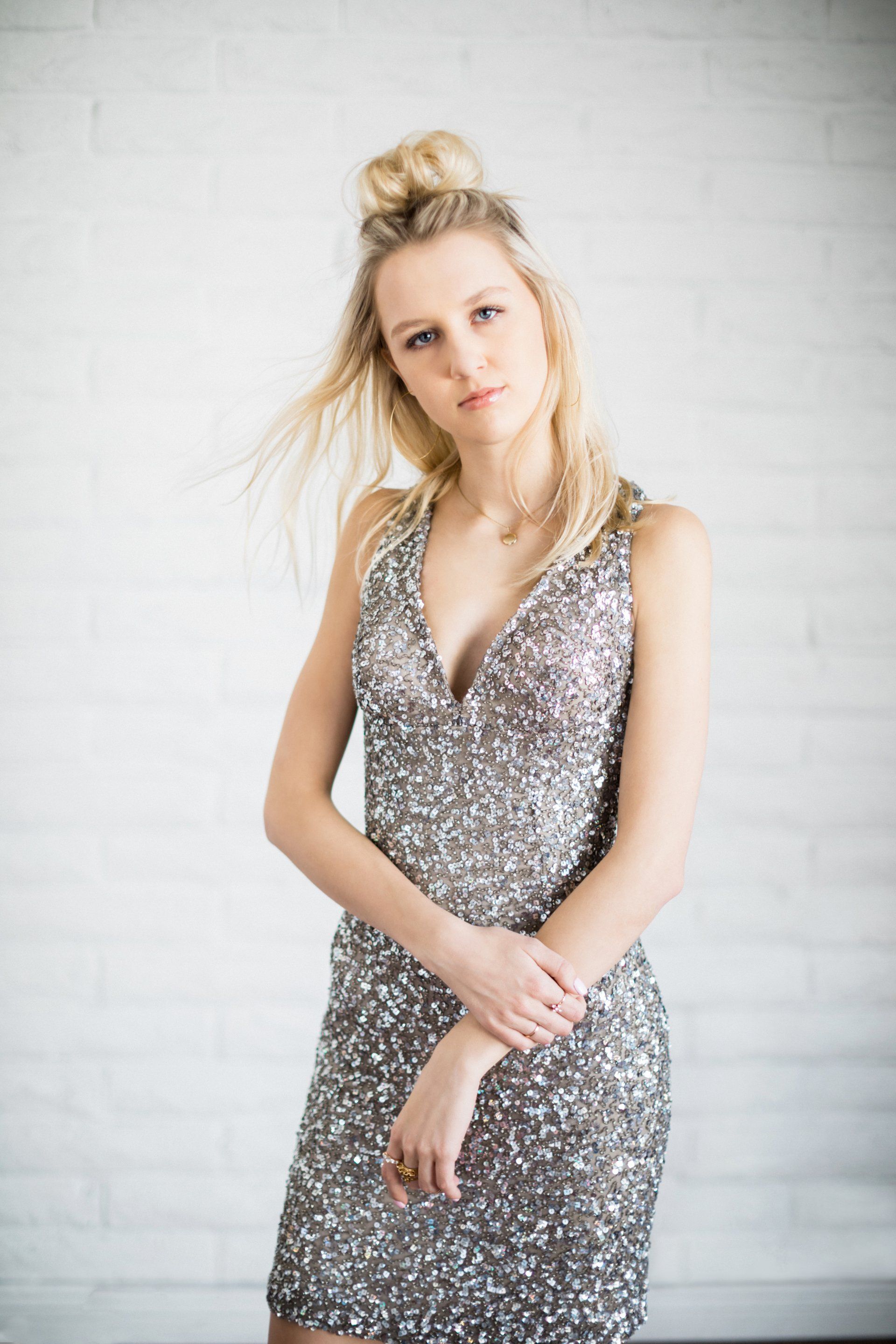 A woman in a silver sequined dress is standing in front of a white brick wall.
