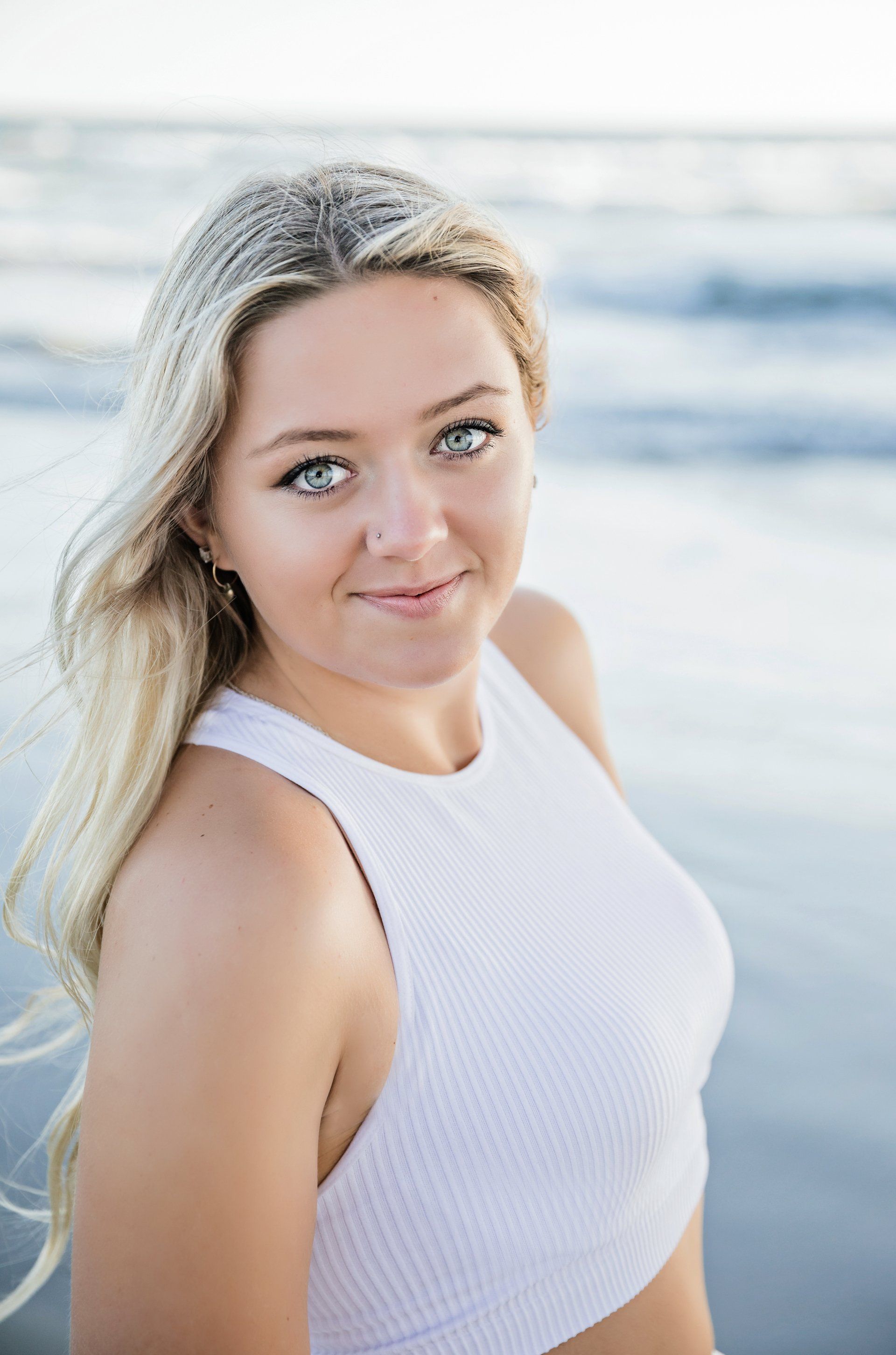 A young woman in a white tank top is sitting on the beach.
