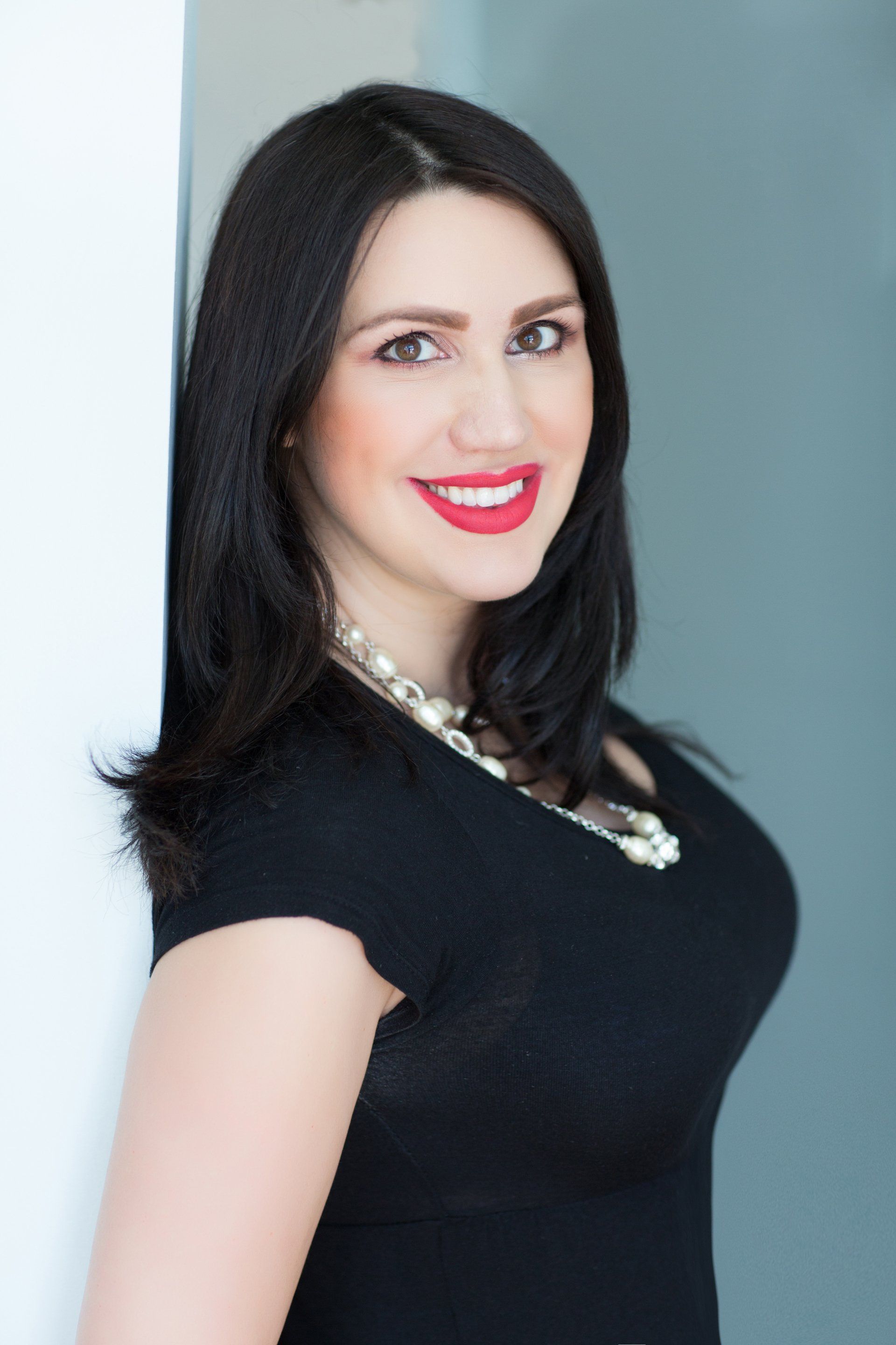 A woman in a black dress is smiling and leaning against a wall