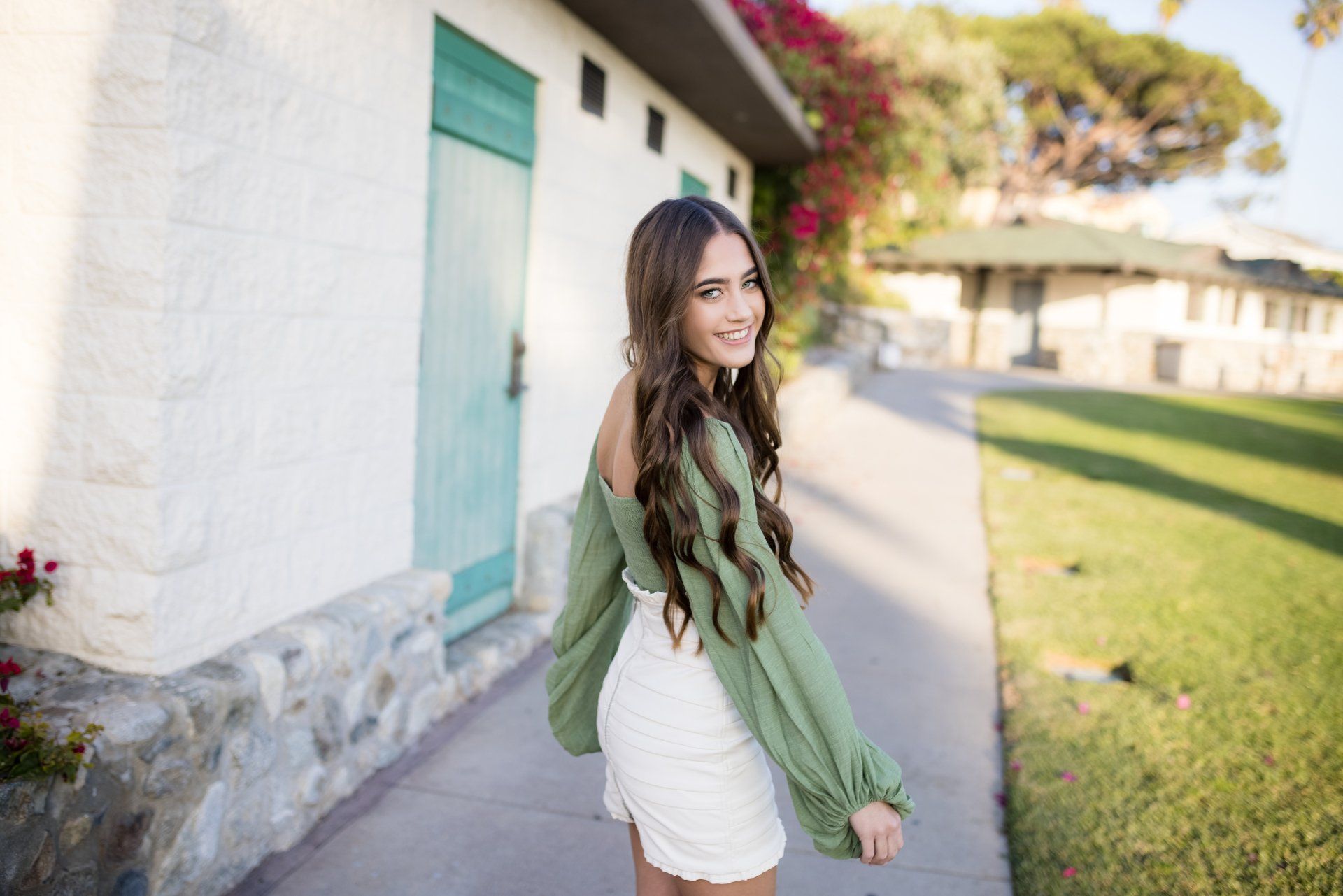 A woman in a green top and white skirt is standing on a sidewalk in front of a building.