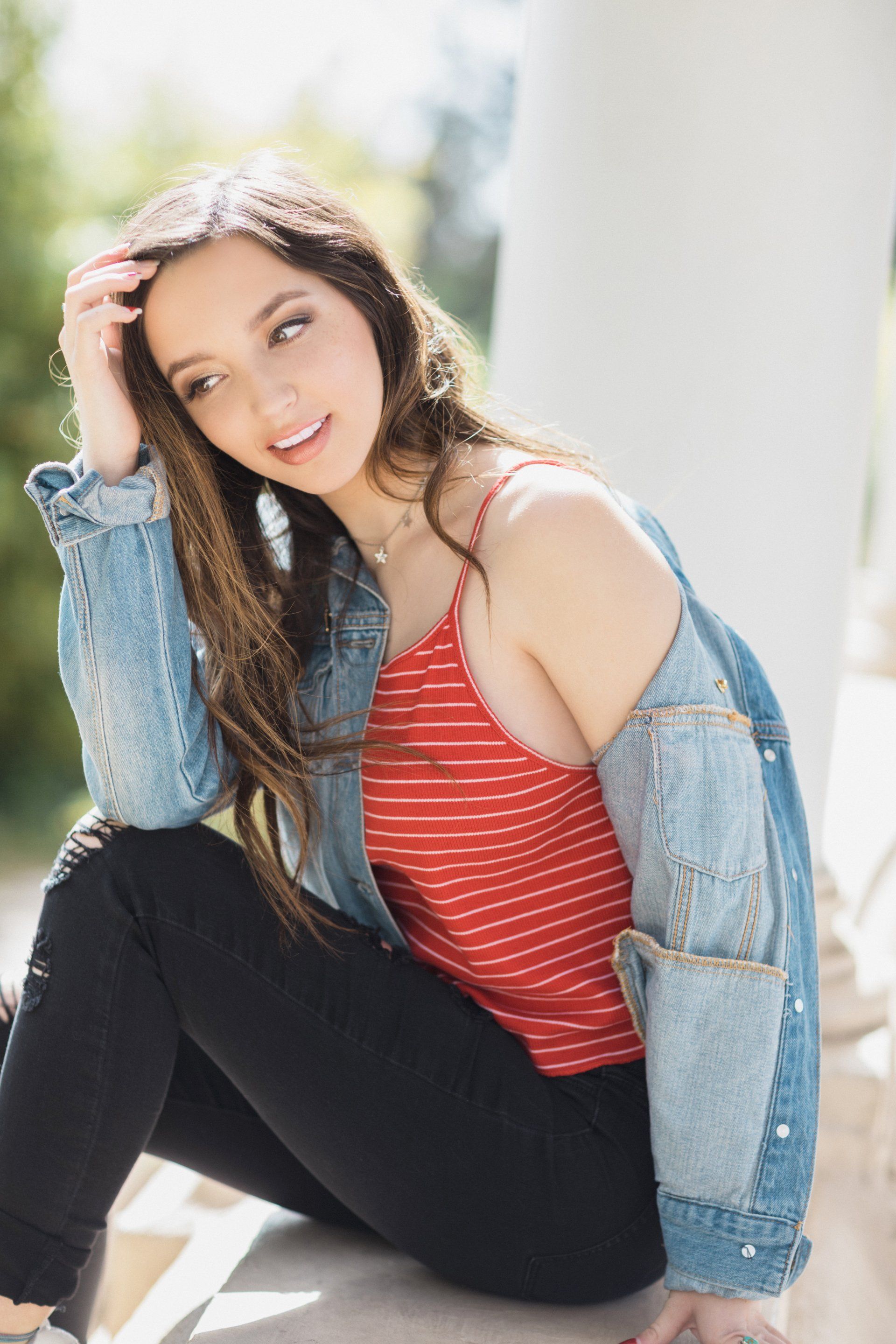 A woman is sitting on the ground wearing a denim jacket and a red tank top.