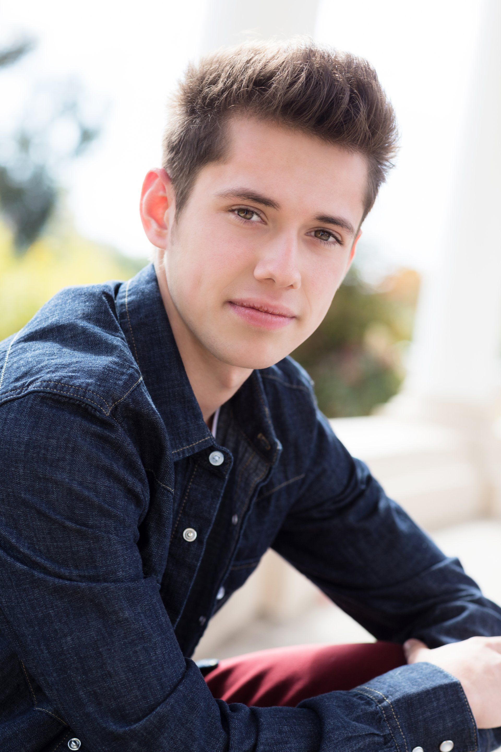 A young man wearing a blue shirt and red pants is sitting down and looking at the camera.