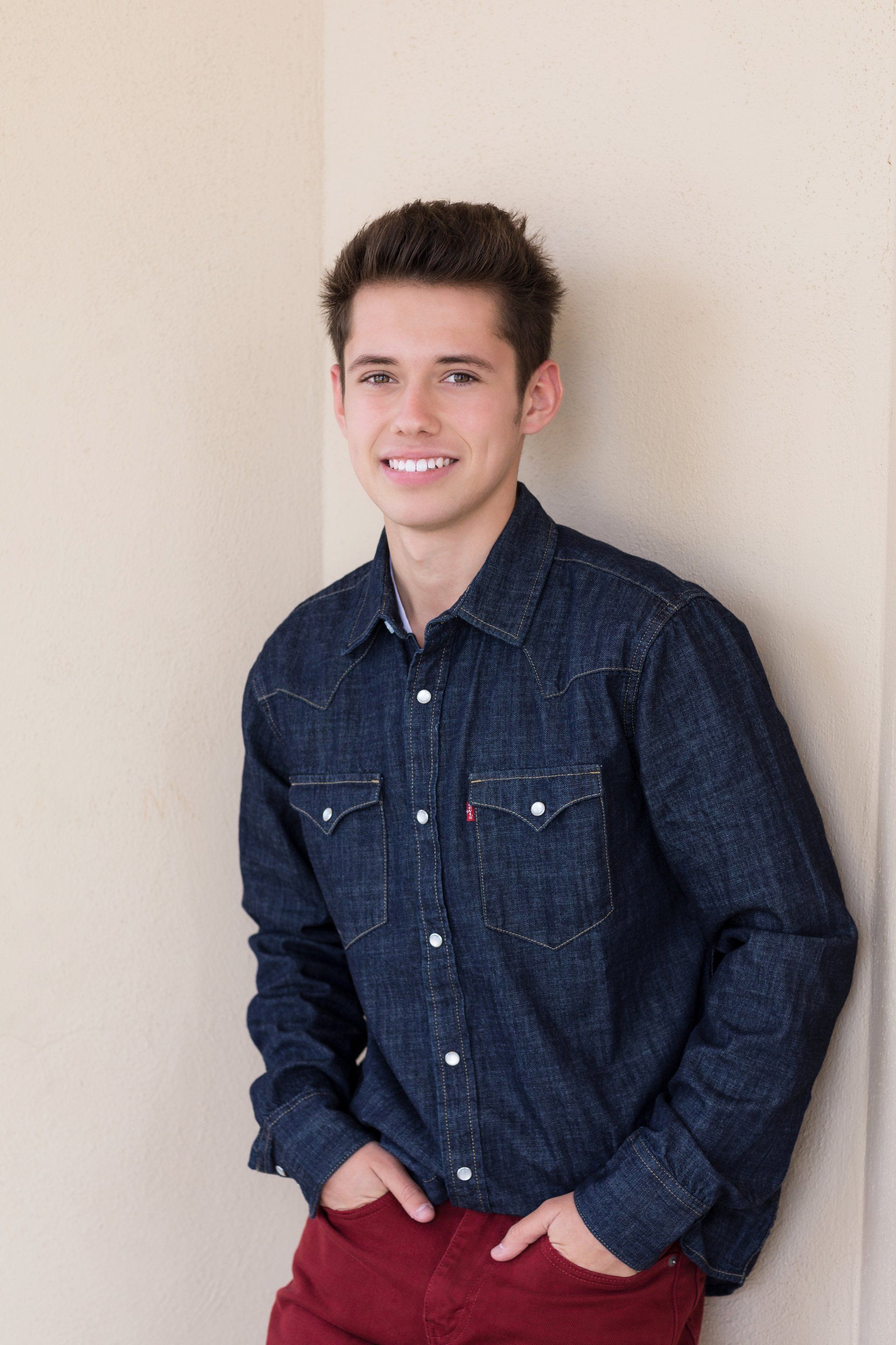 A young man in a denim shirt and red pants is leaning against a wall.