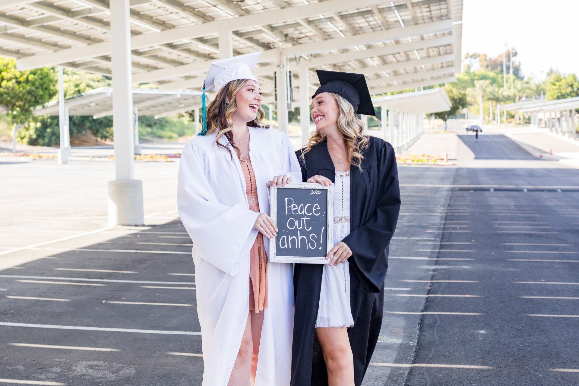 Two female graduates are standing next to each other in a parking lot holding a sign.