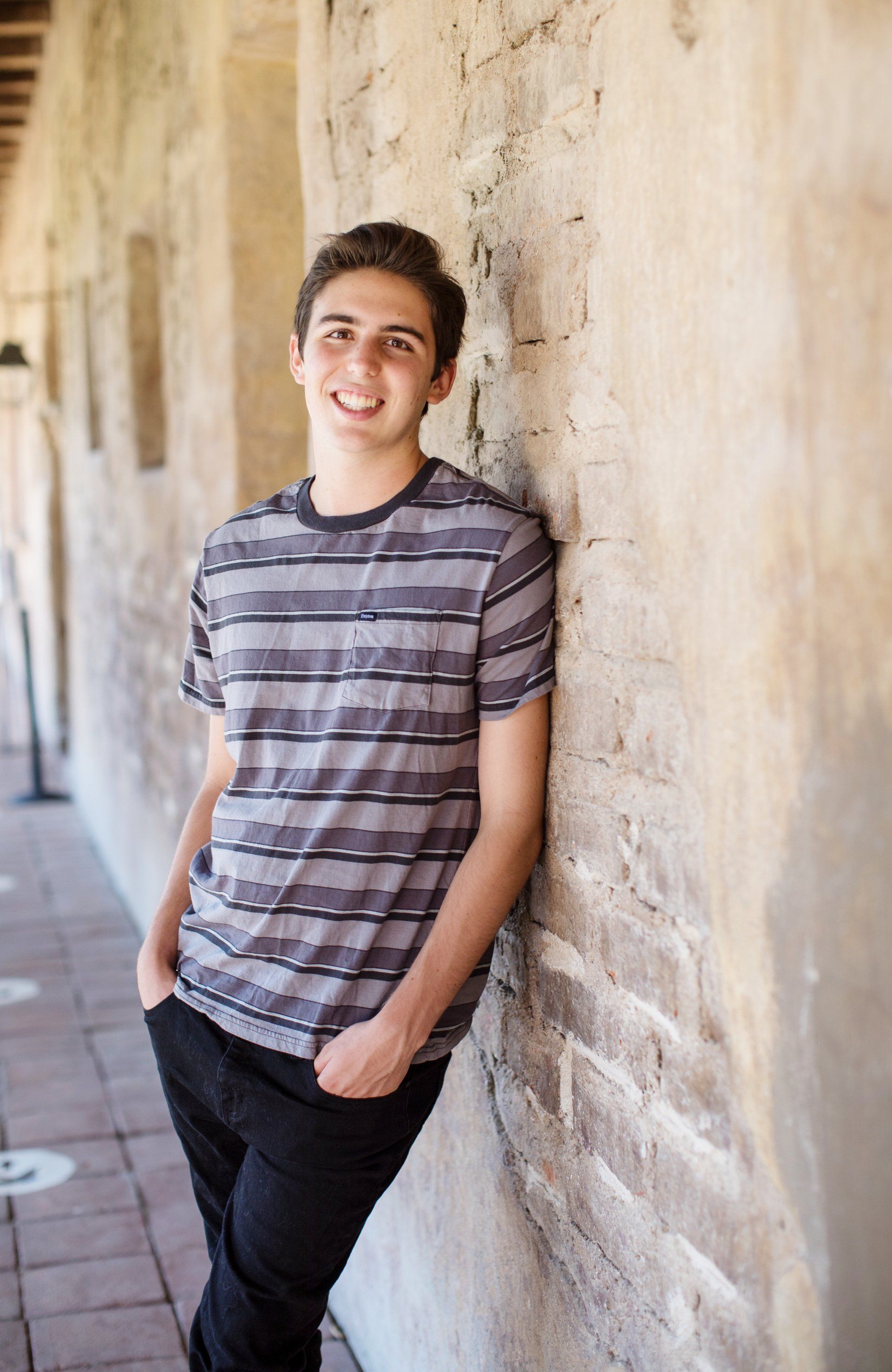 A young man leaning against a brick wall with his hands in his pockets.