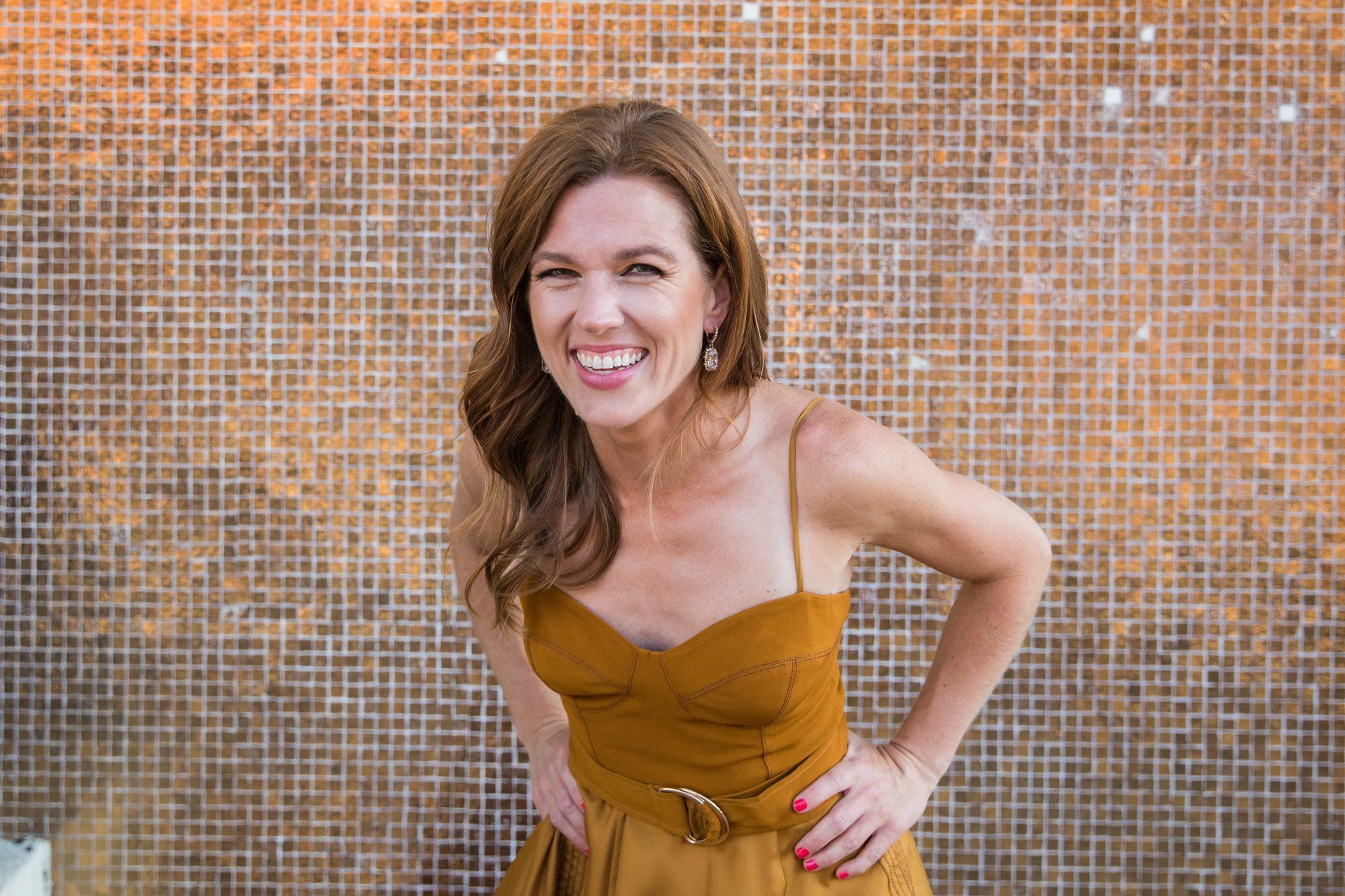 A woman in a yellow dress is standing in front of a tiled wall and smiling.