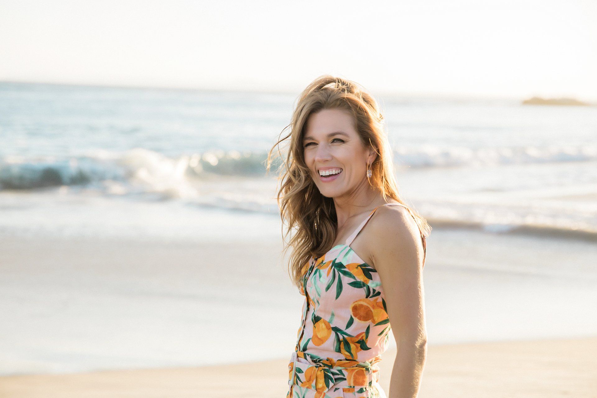 A woman in a floral dress is standing on the beach and smiling.