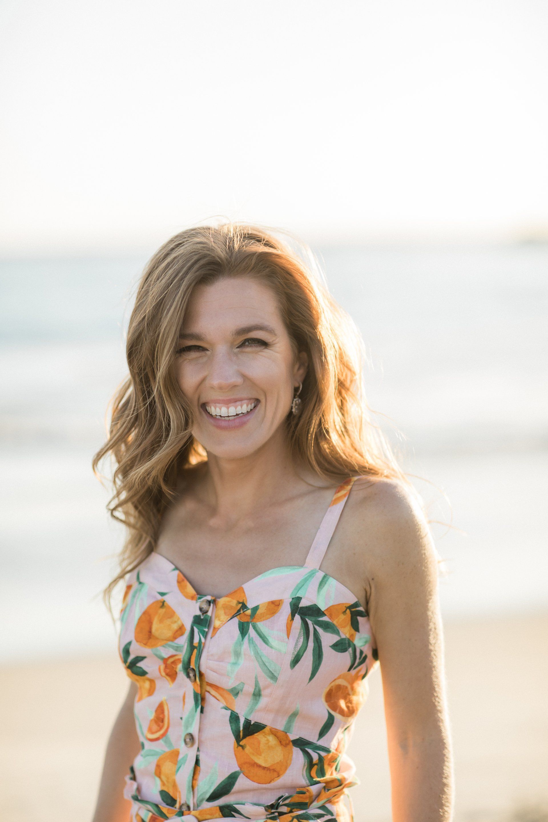 A woman in an orange floral dress is standing on the beach and smiling.