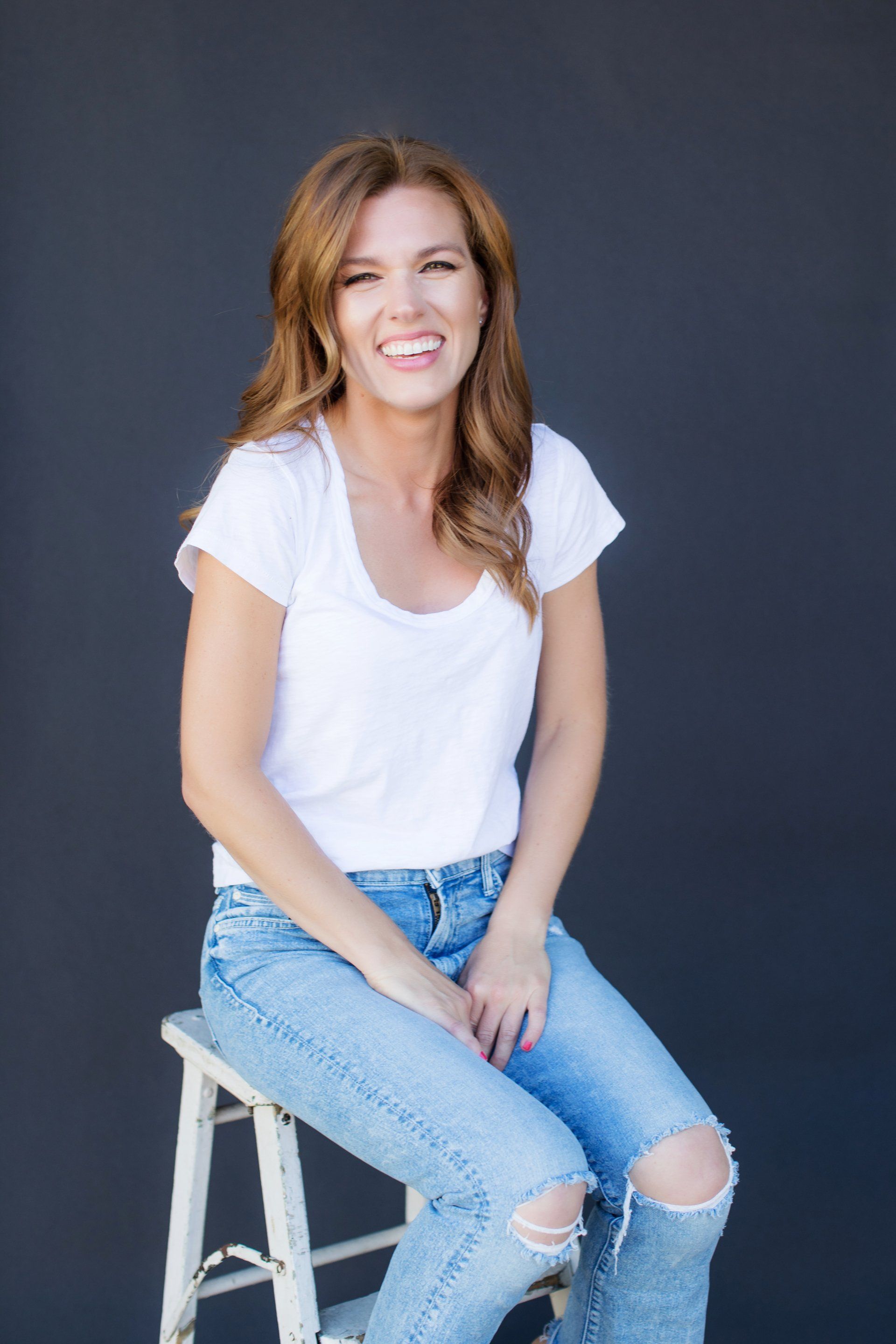 A woman in a white shirt and ripped jeans is sitting on a stool.