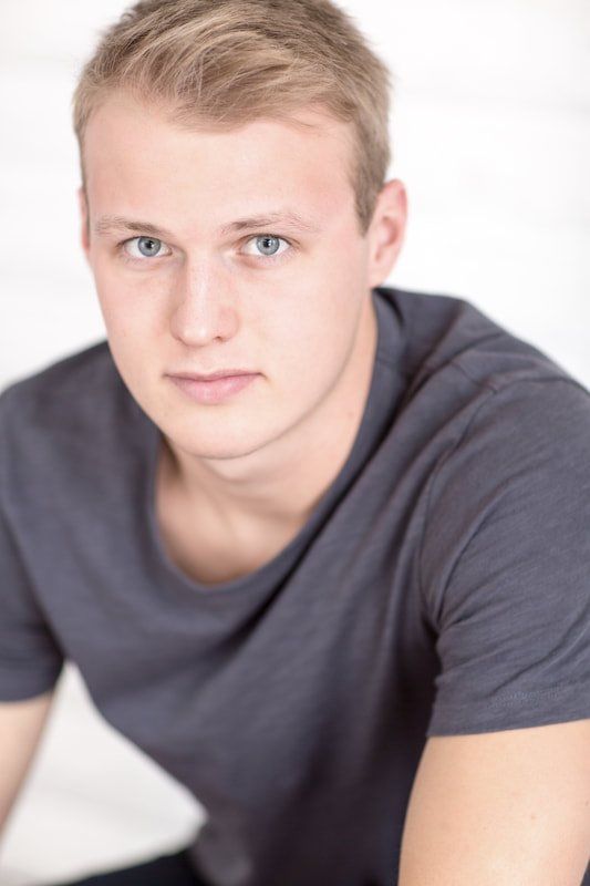 A young man wearing a grey t-shirt is sitting down and looking at the camera.