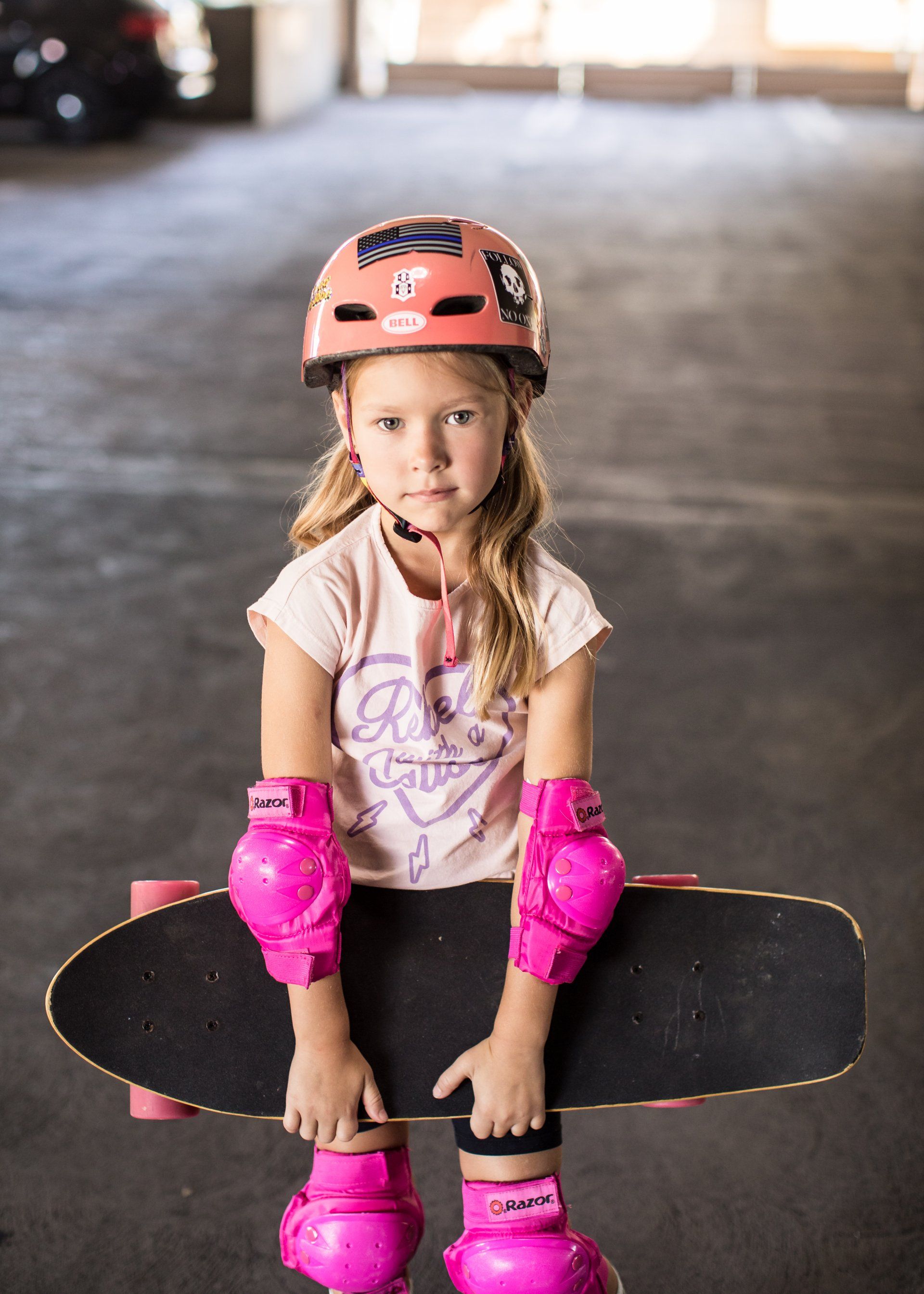 A little girl wearing a helmet and pink knee pads is sitting on a skateboard.