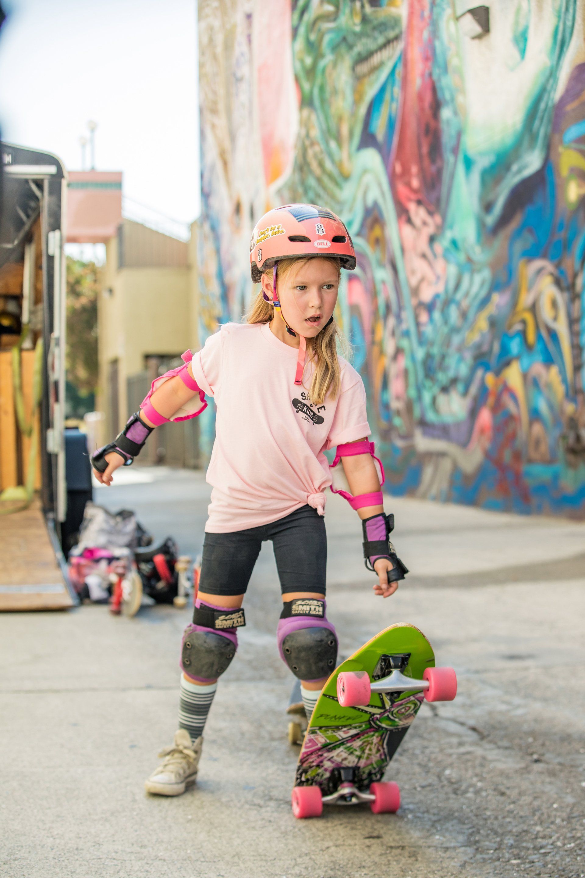 A young girl is riding a skateboard on a sidewalk in front of a graffiti wall.