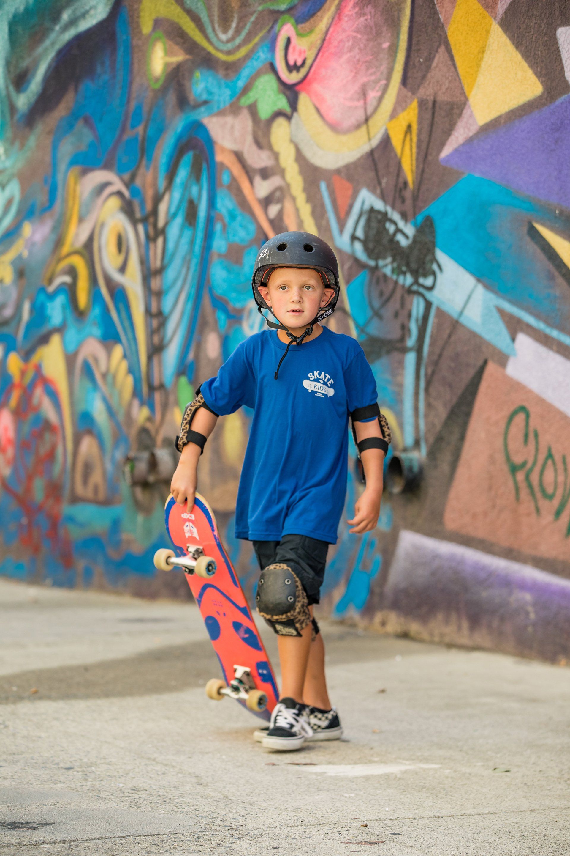 A young boy is holding a skateboard in front of a graffiti wall.