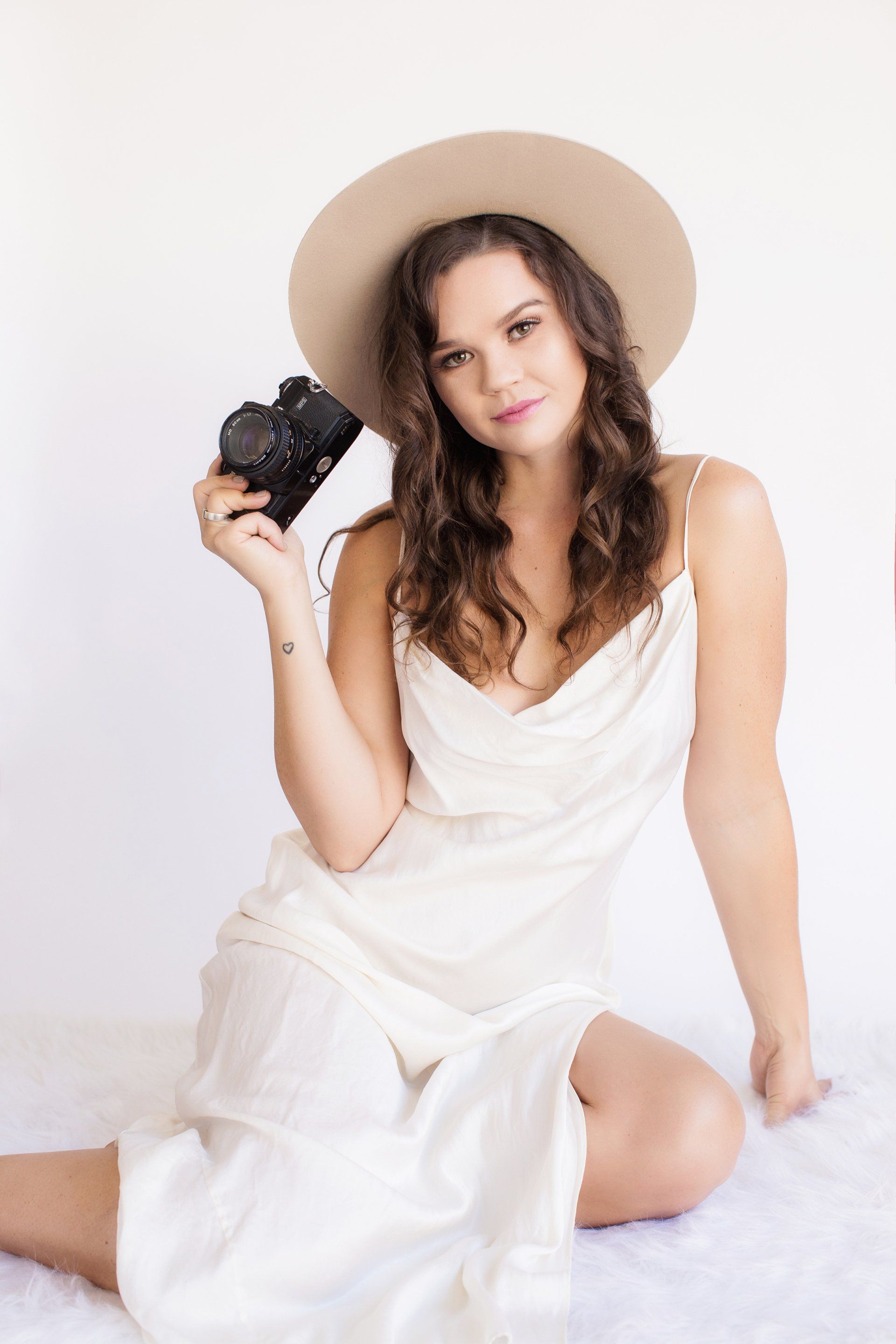 A woman in a white dress and hat is sitting on the floor holding a camera.