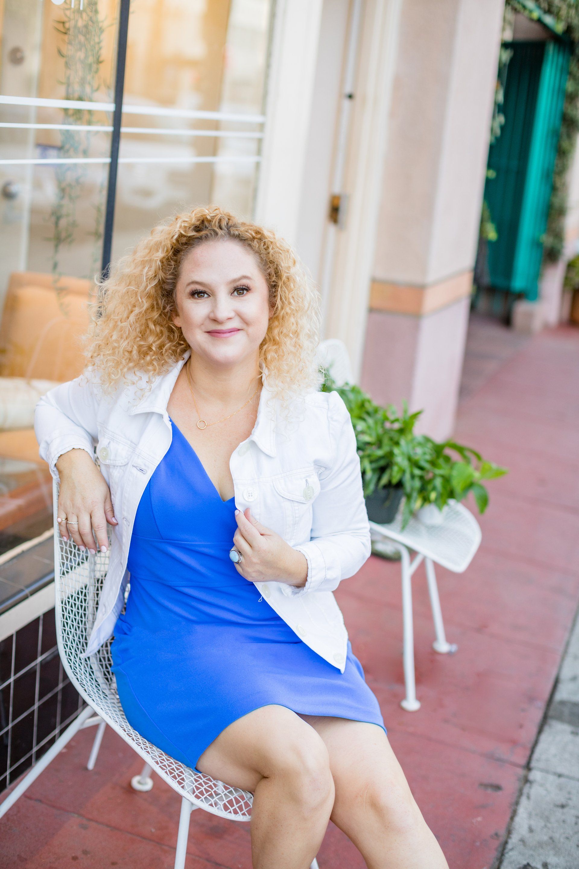 A woman in a blue dress and white jacket is sitting in a chair on the sidewalk.