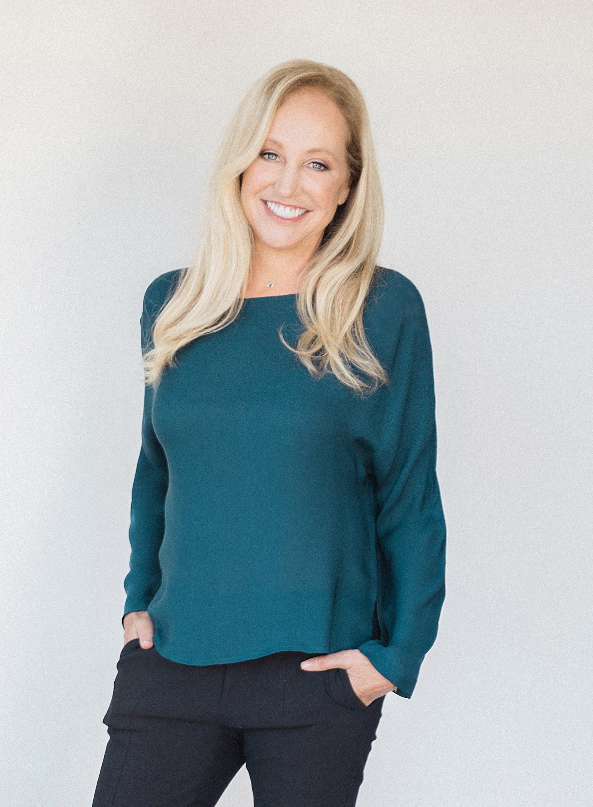 A woman wearing a teal sweater and black pants is standing with her hands in her pockets.