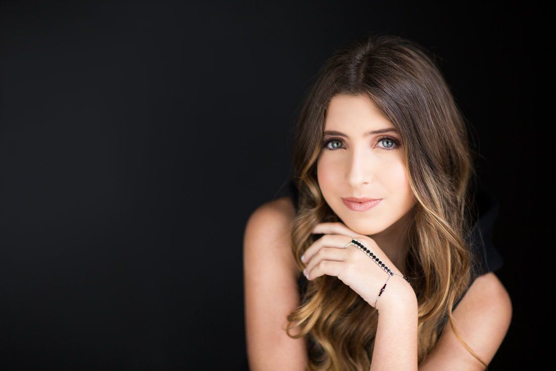 A woman with long hair is sitting in front of a black background with her hand on her chin.