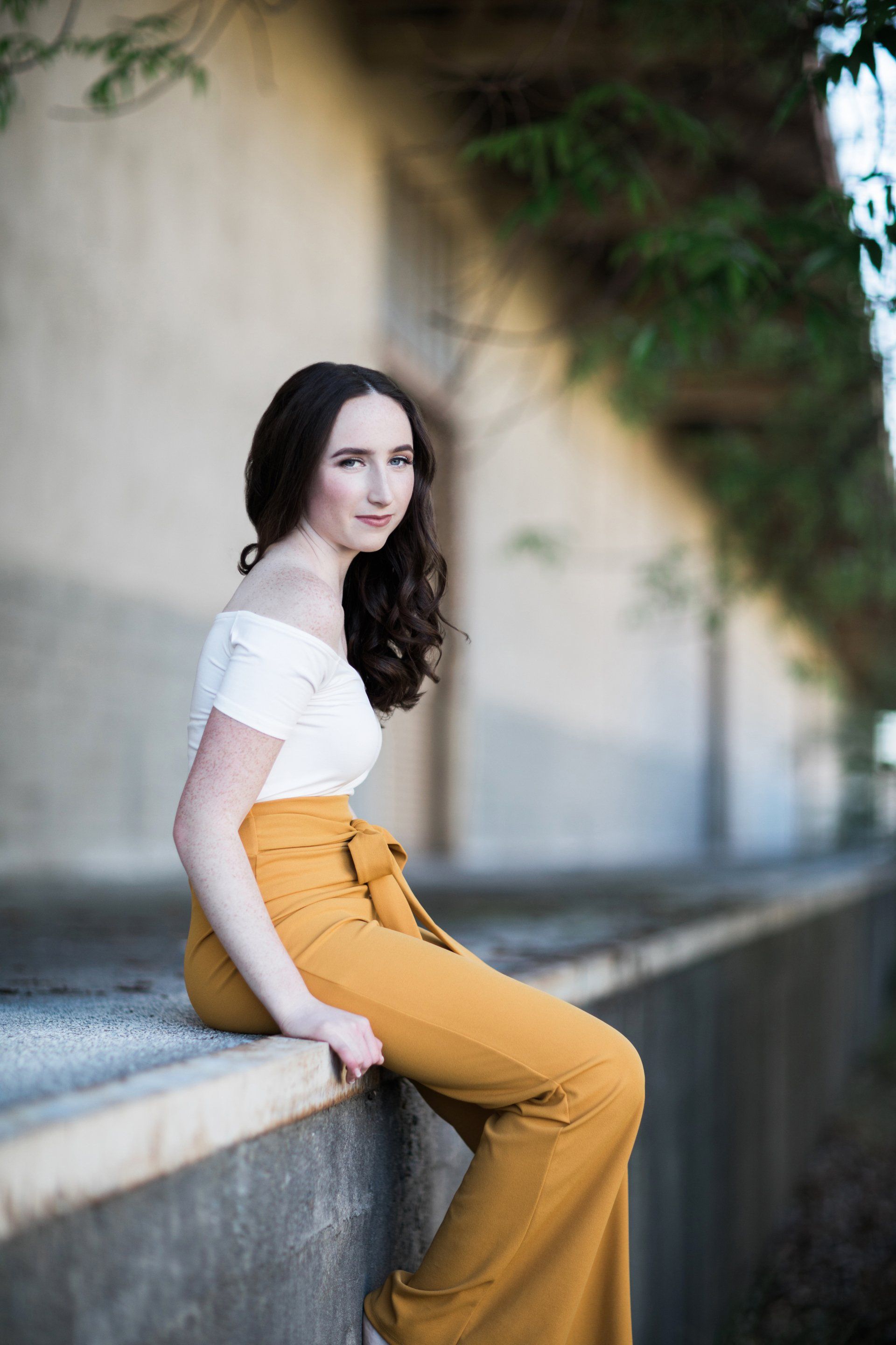 A woman is sitting on a wall wearing yellow pants and a white top.