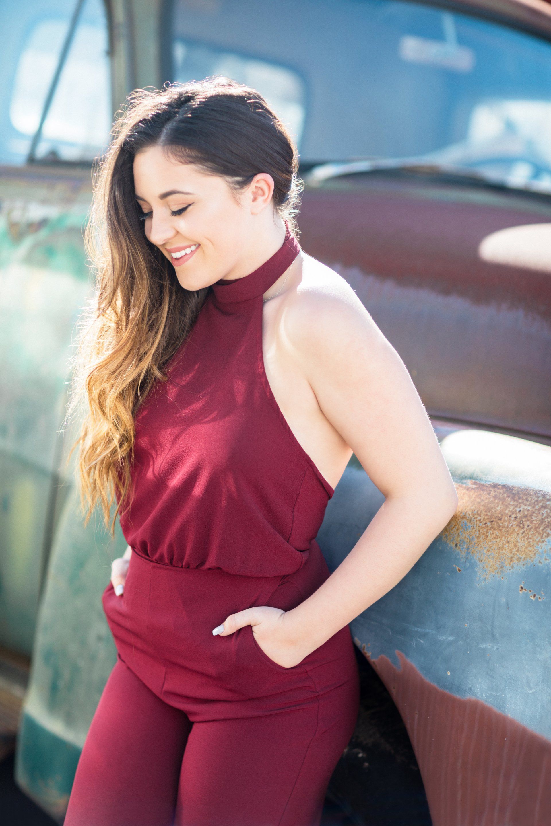 A woman in a red jumpsuit is leaning against a rusty truck.