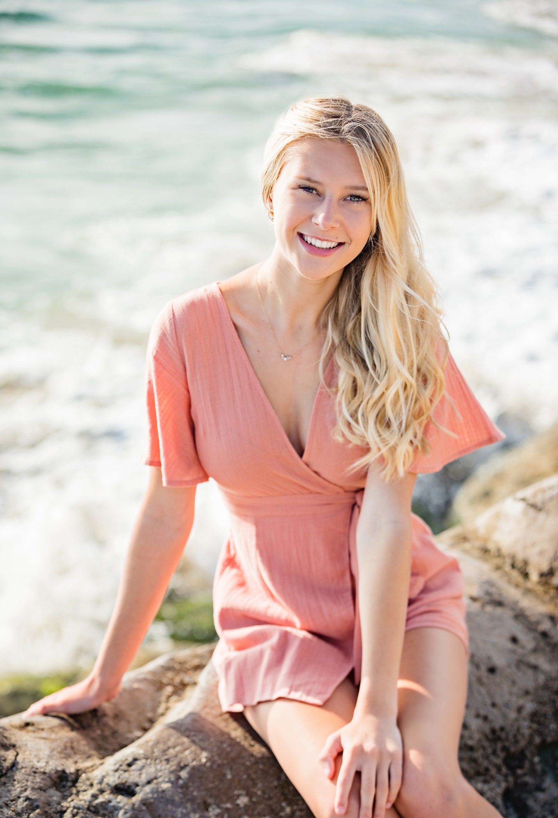 A woman in a pink dress is sitting on a rock near the ocean.