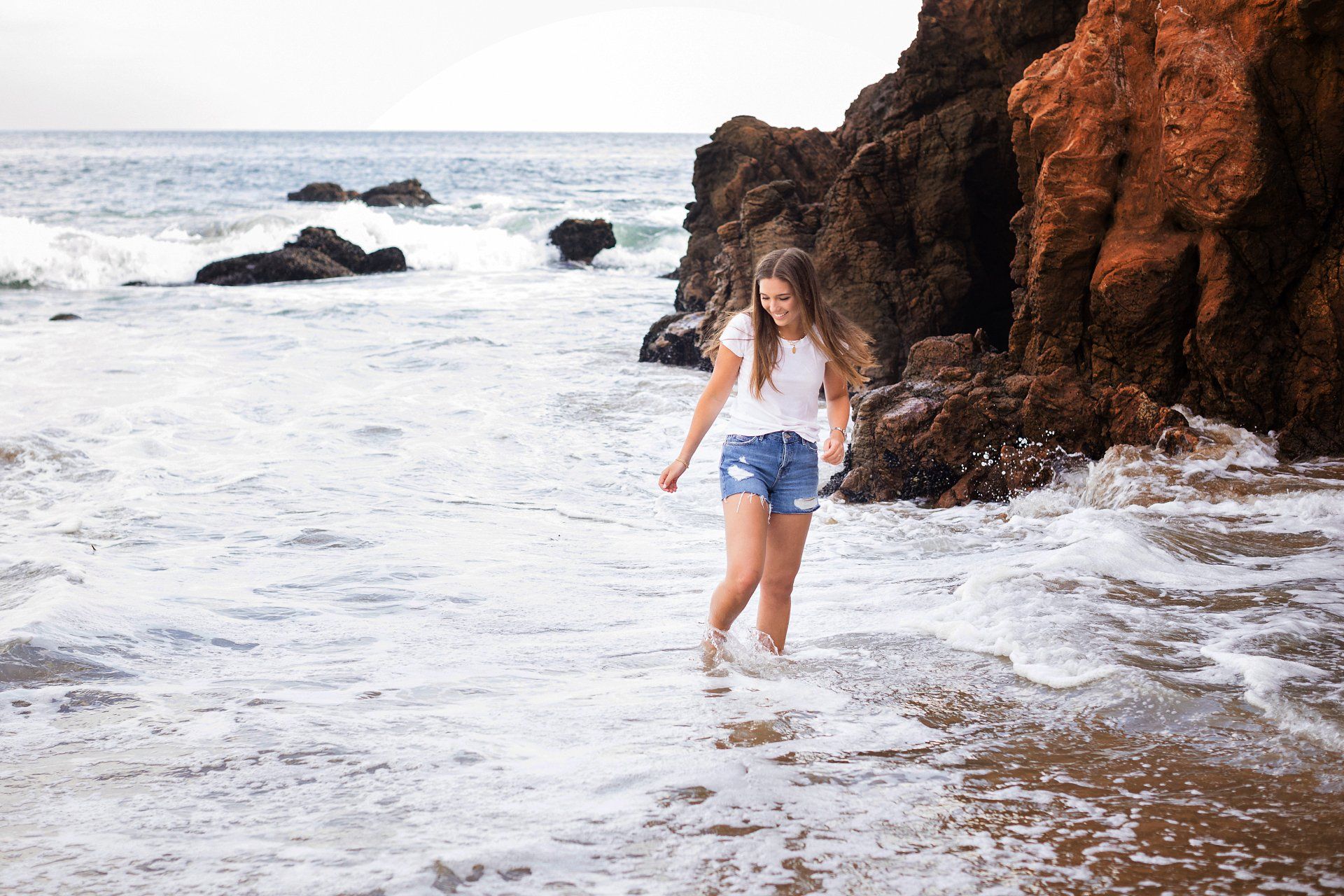 A woman is walking into the ocean on a rocky beach.