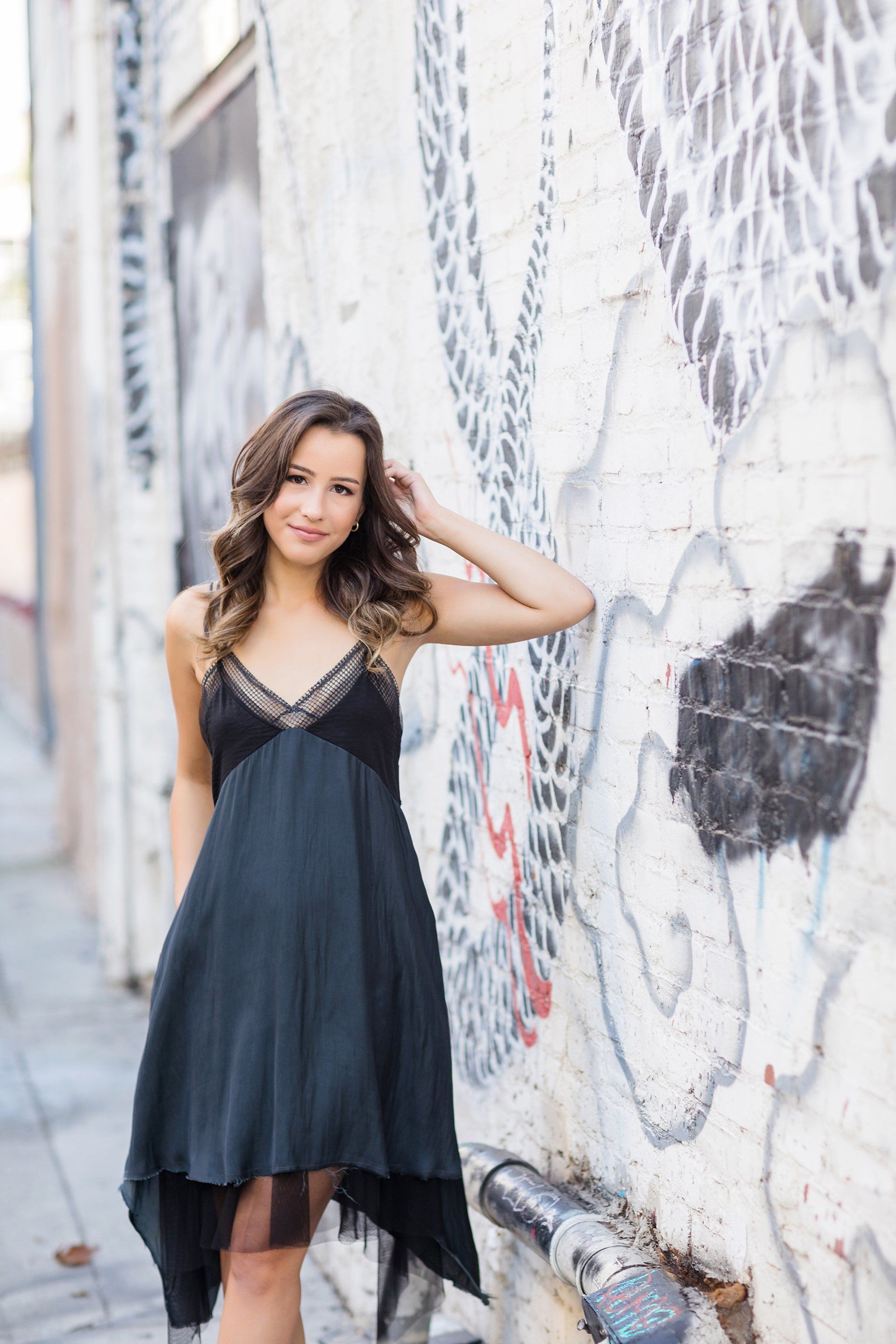 A woman in a black dress is standing in front of a graffiti covered wall.