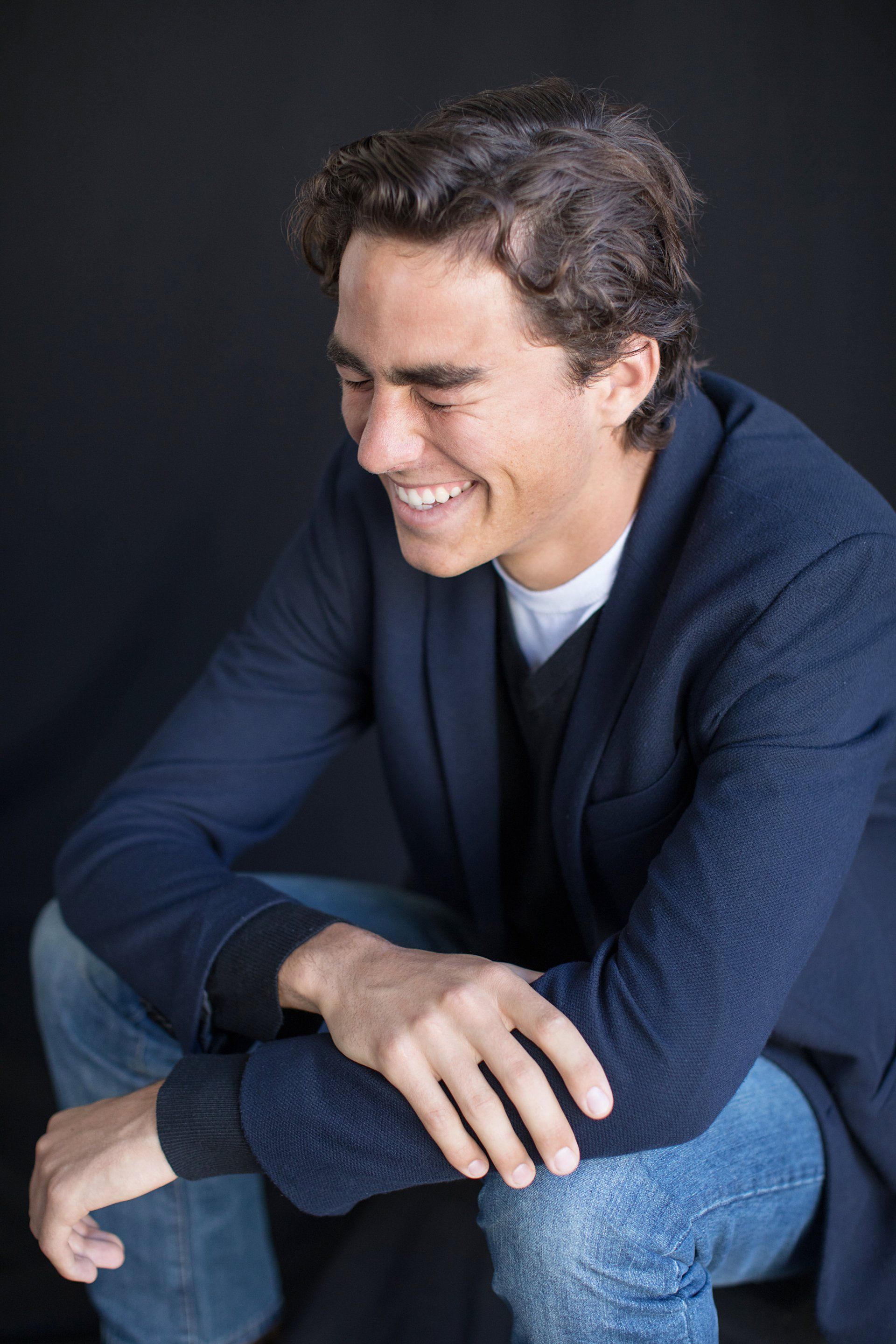 A man in a blue jacket is sitting down with his arms crossed and smiling.