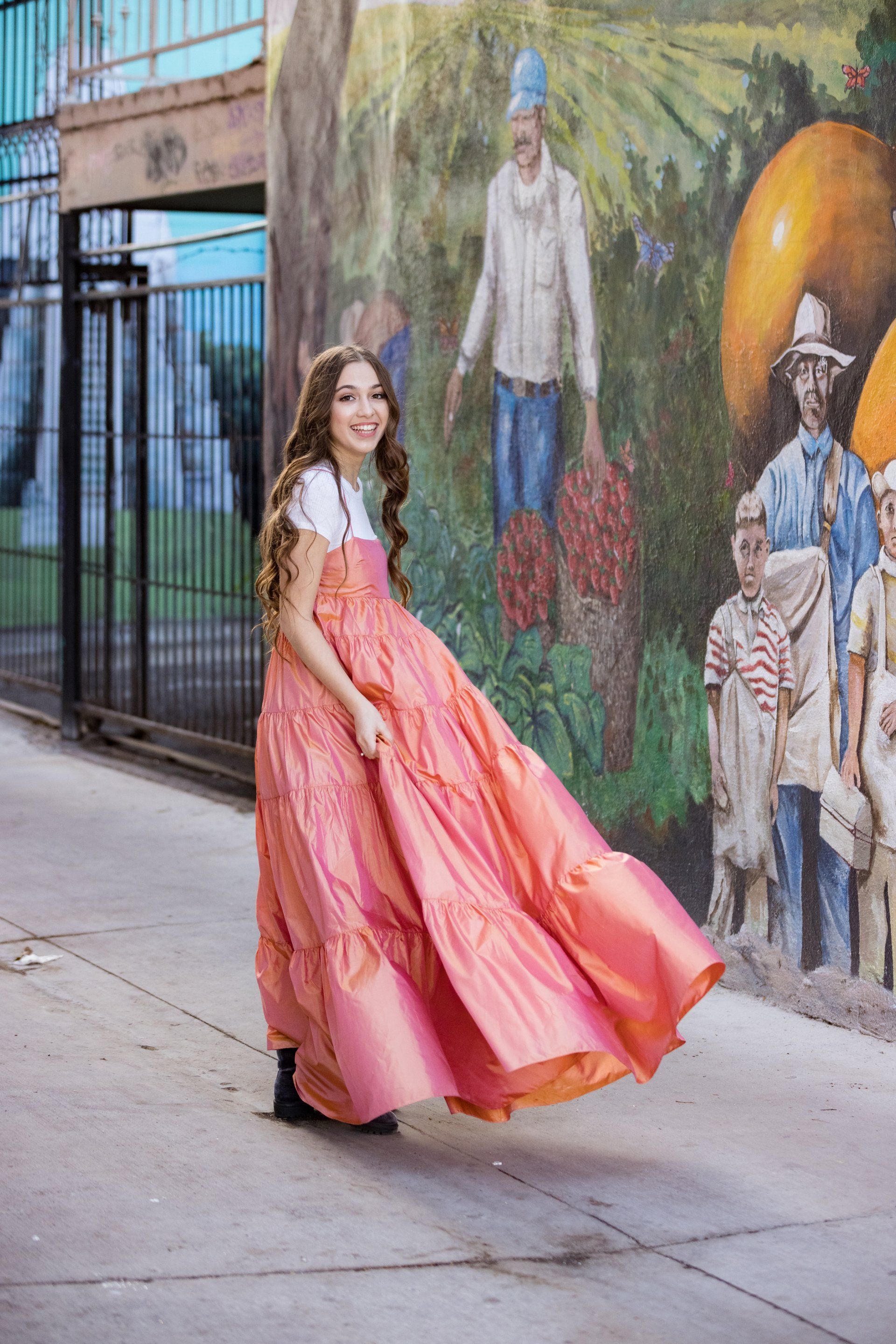 A woman in a long pink dress is standing on a sidewalk in front of a mural.