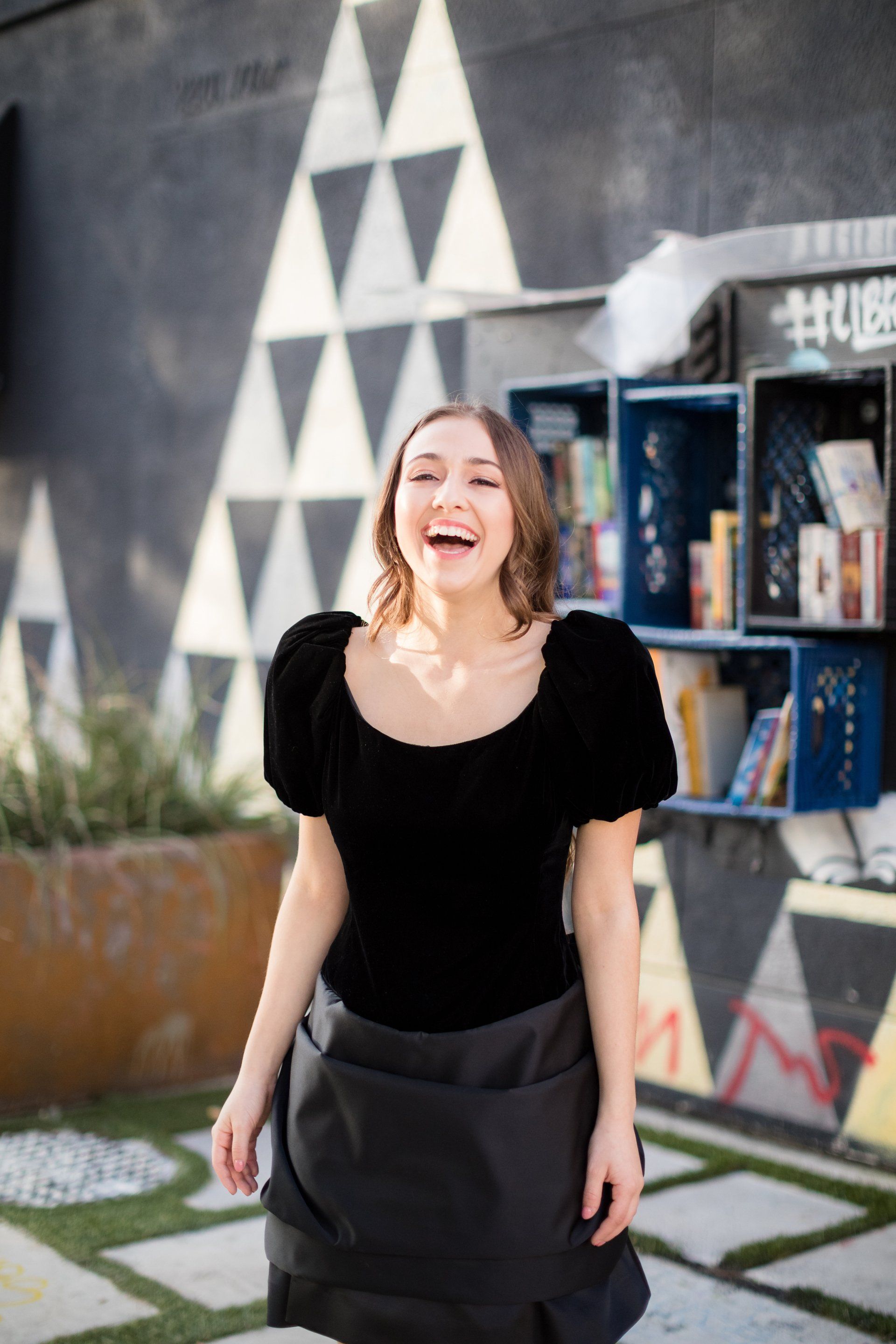 A woman in a black top and black skirt is standing in front of a bookshelf.