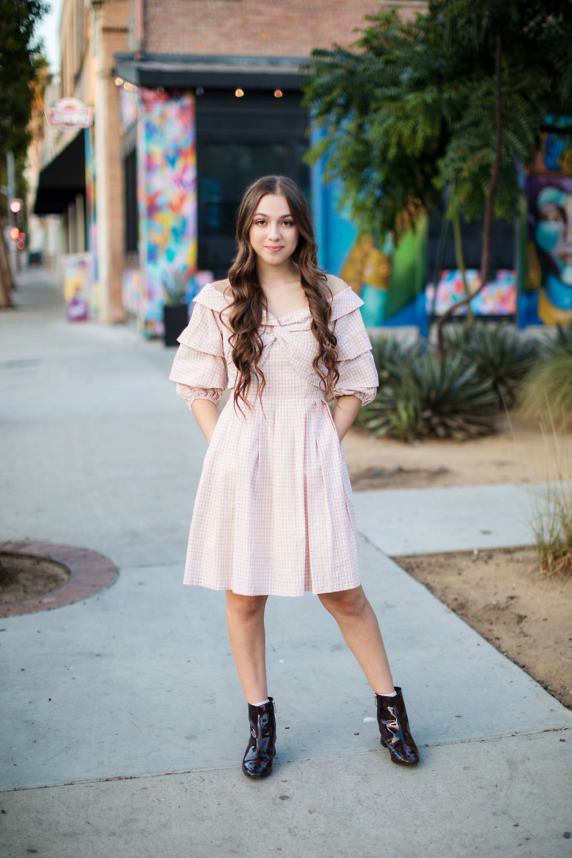 A young woman in a pink dress and black boots is standing on a sidewalk.
