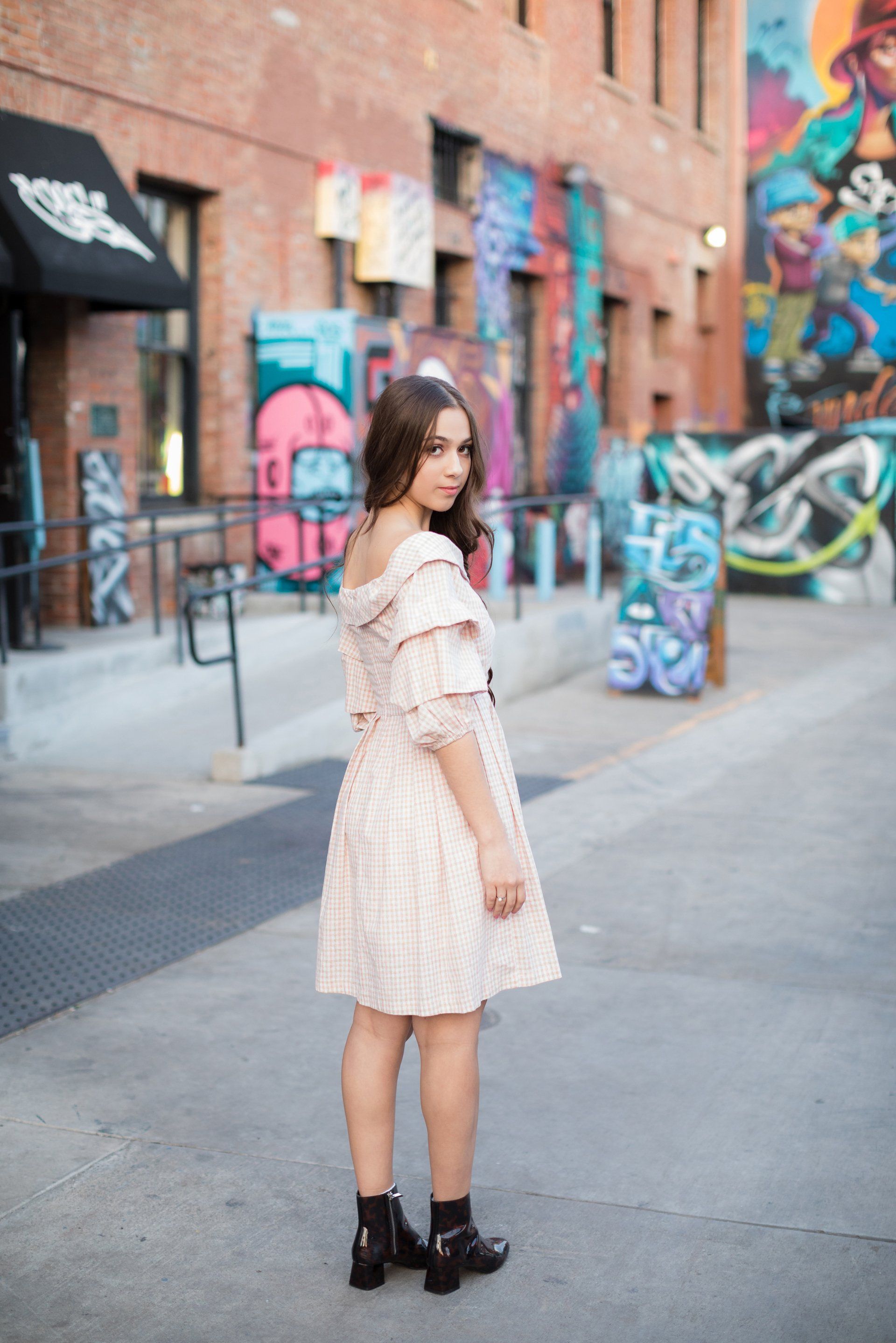 A woman in a pink dress is standing in front of a brick building with graffiti on it.