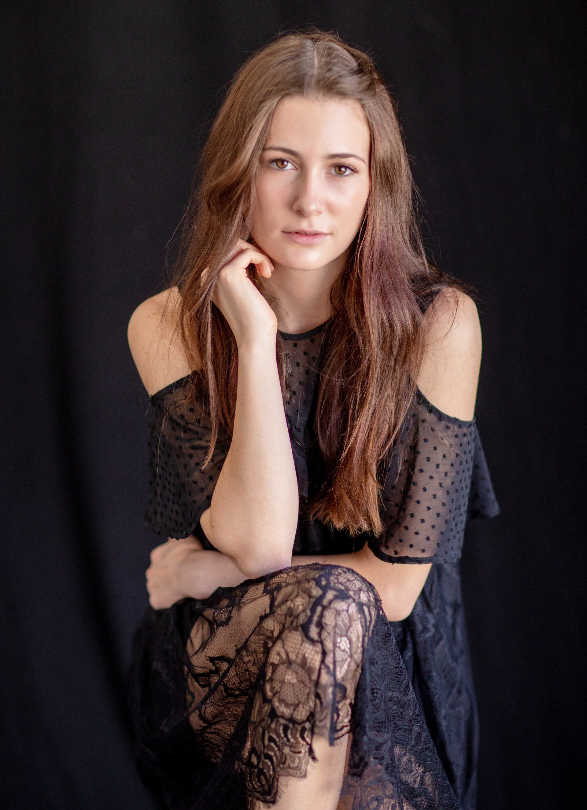 A woman in a black dress is sitting with her legs crossed and her hand on her chin.