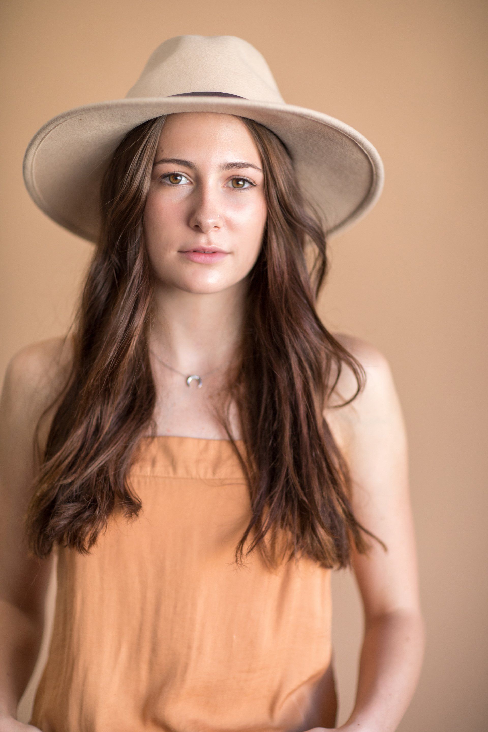 A woman wearing a hat and an orange top is standing in front of a brown background.