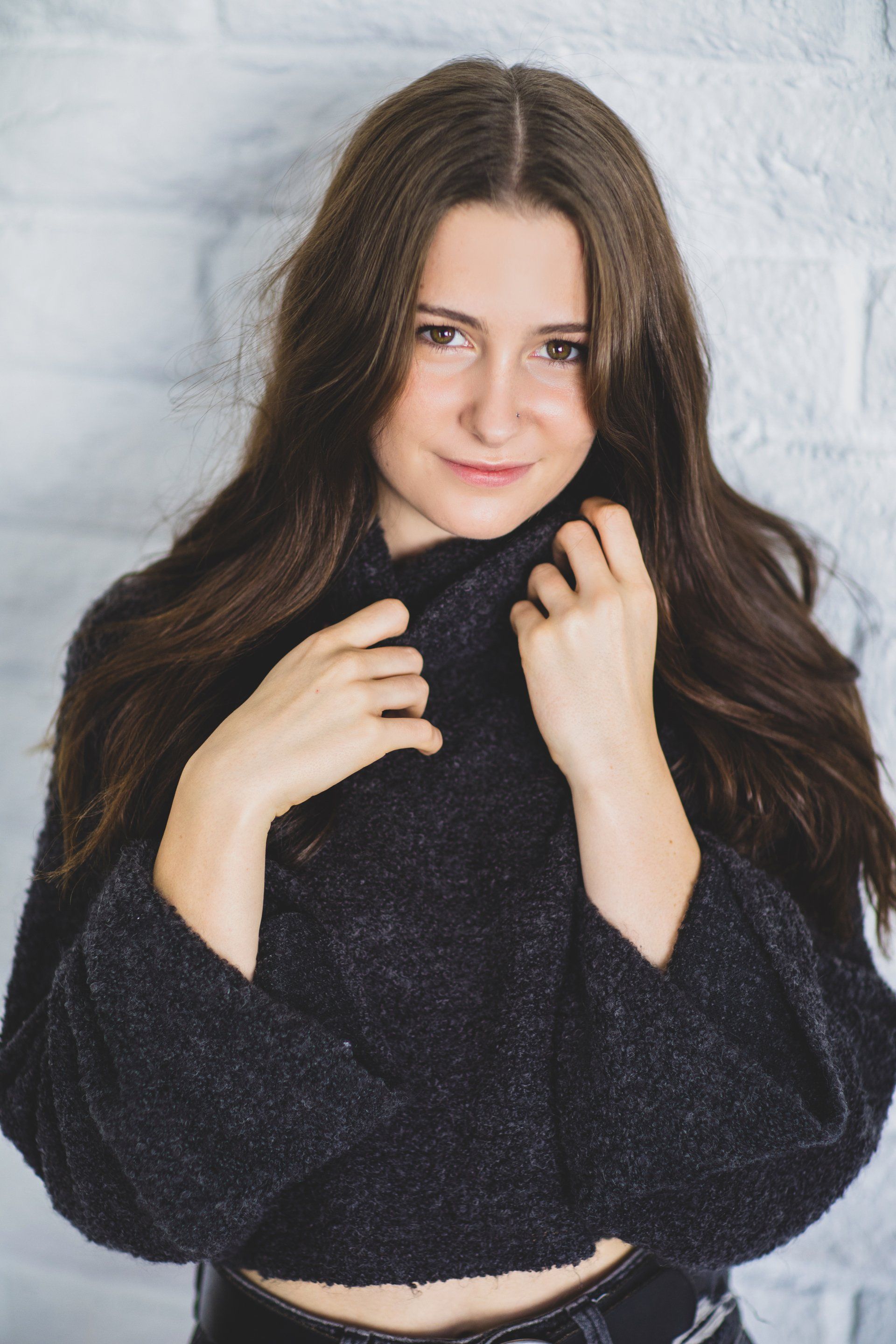 A woman wearing a black sweater is standing in front of a white brick wall.