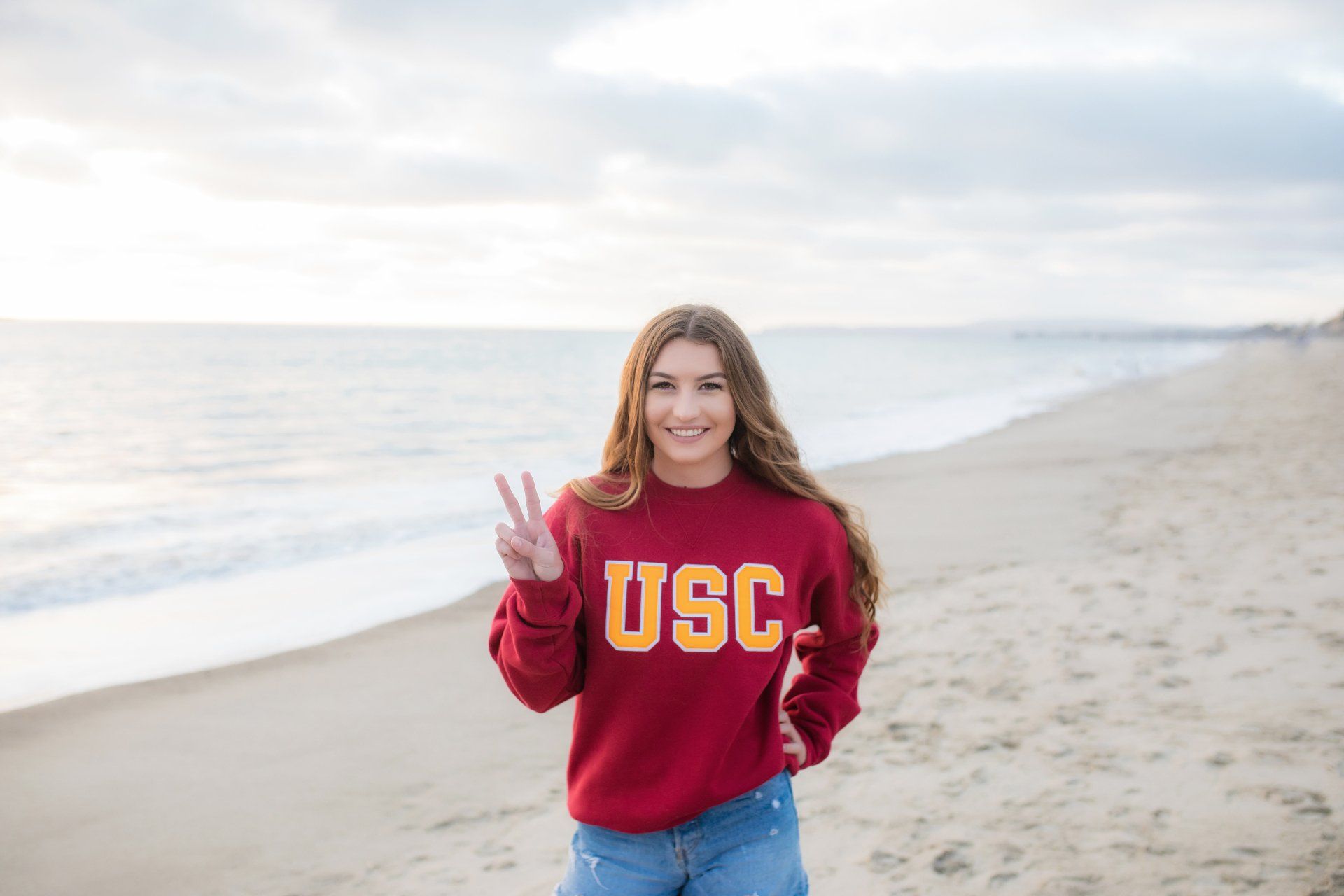 A woman in a usc sweatshirt is standing on a beach giving a peace sign.