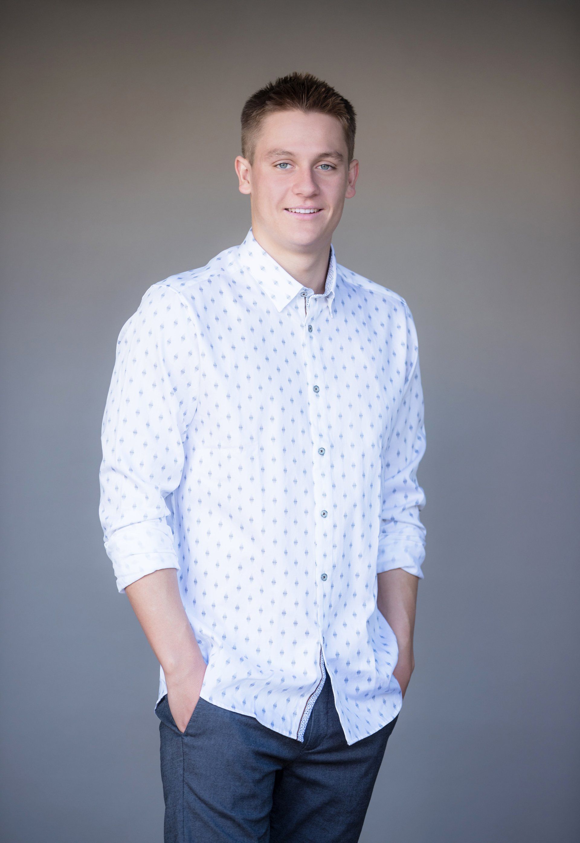 A young man wearing a white shirt and gray pants is standing with his hands in his pockets.