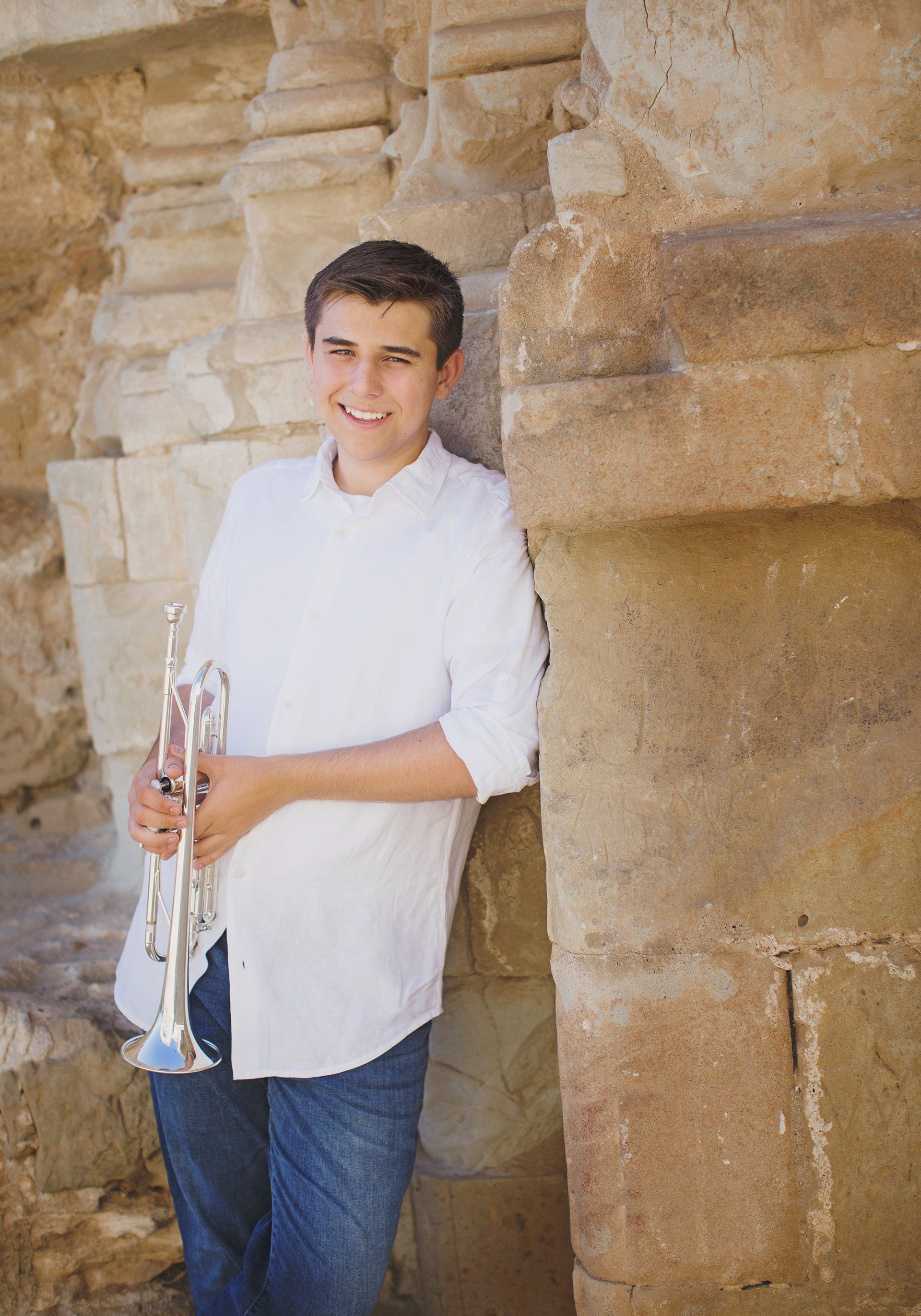 A young man is holding a trumpet while leaning against a stone wall.