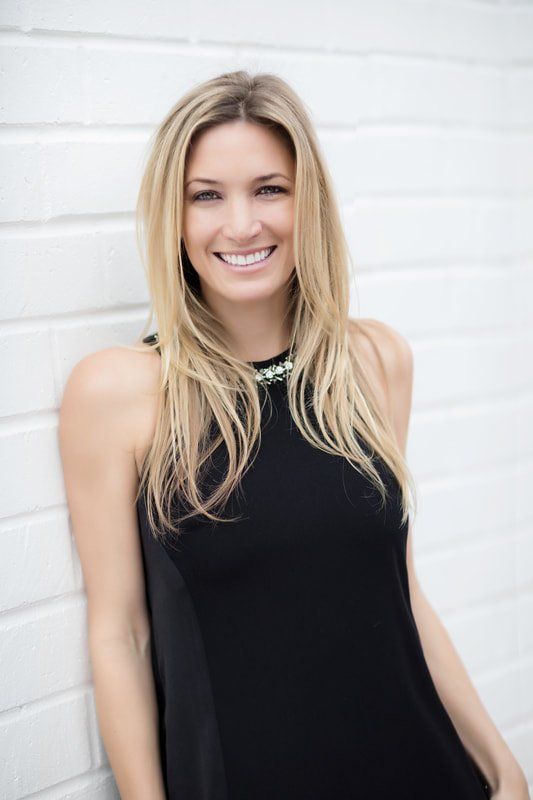 A woman in a black dress is standing in front of a white wall and smiling.