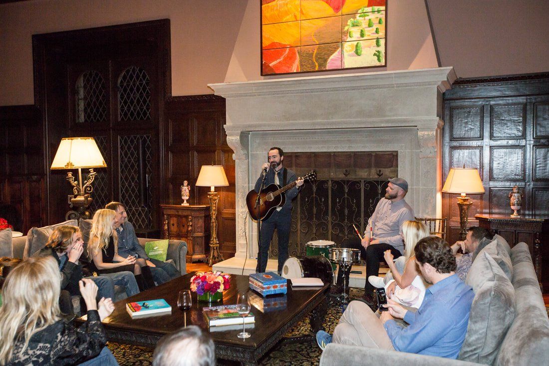 A man is playing a guitar in front of a group of people in a living room.