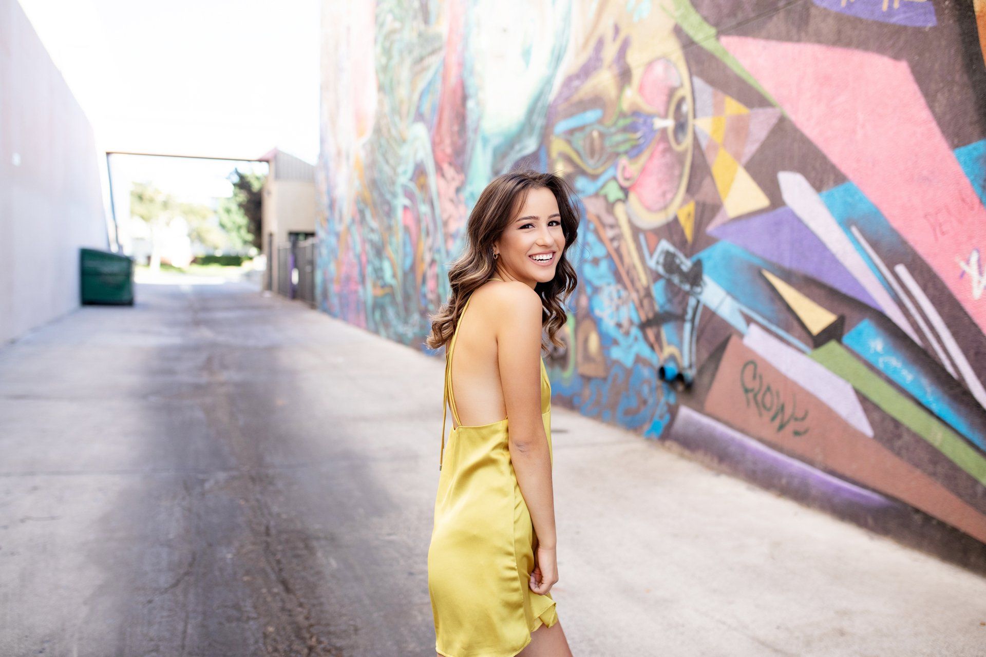 A woman in a yellow dress is standing in front of a graffiti wall.
