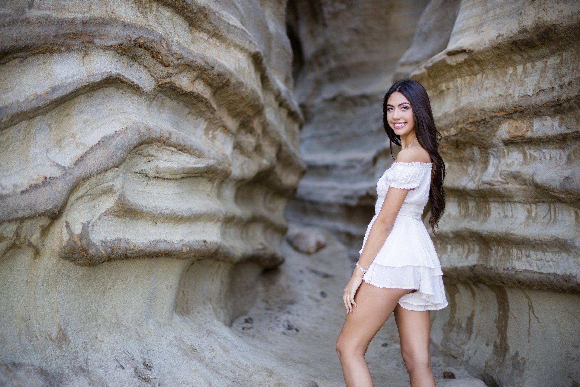 A woman in a white dress is standing in front of a rock wall.