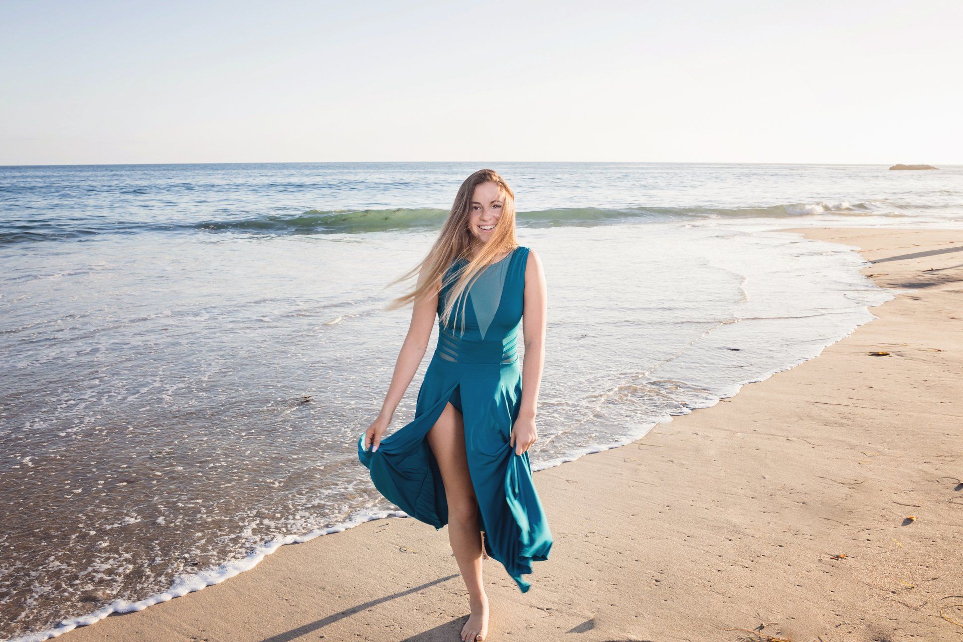 A woman in a blue dress is standing on the beach near the ocean.