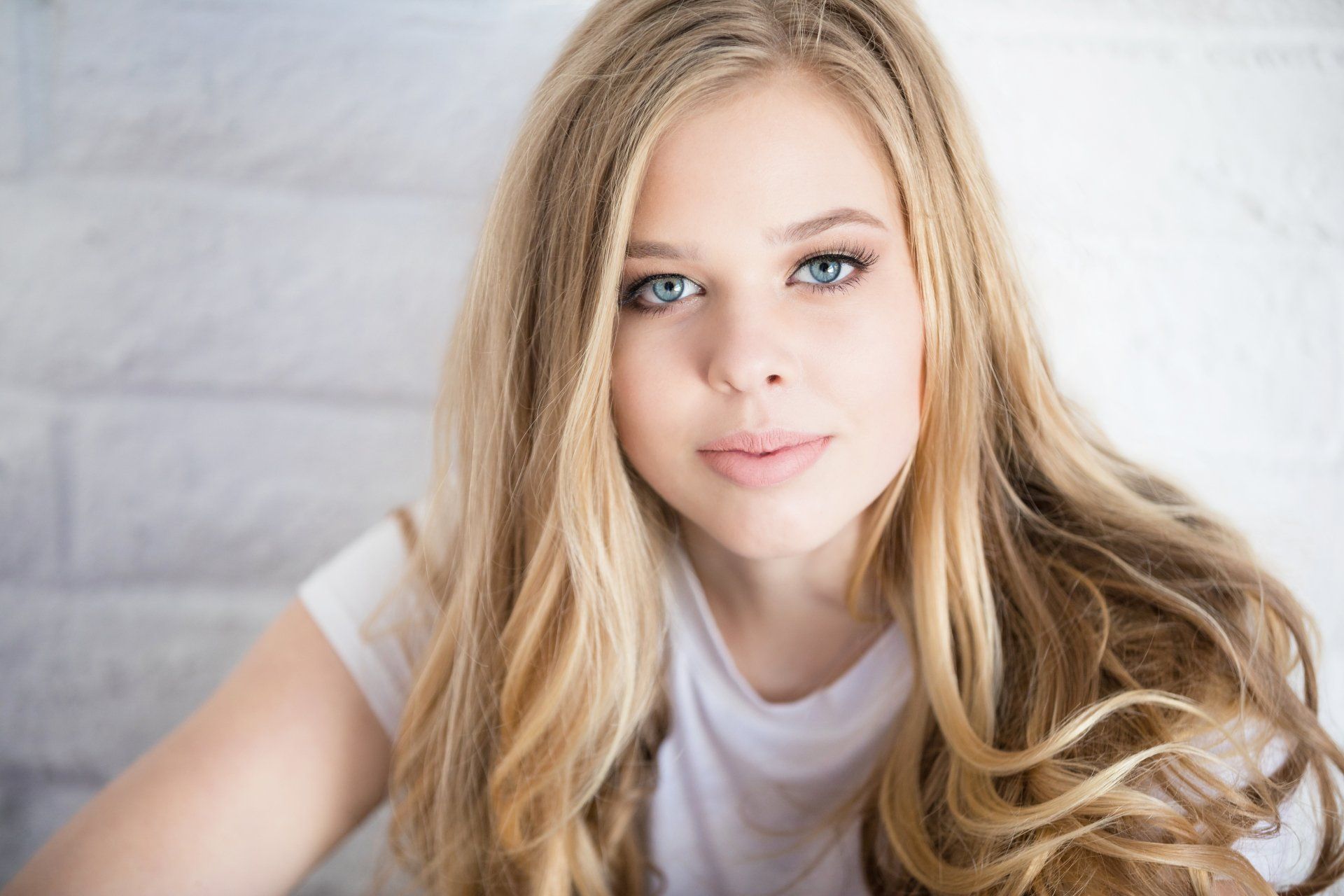 A young woman with long blonde hair and blue eyes is wearing a white shirt.