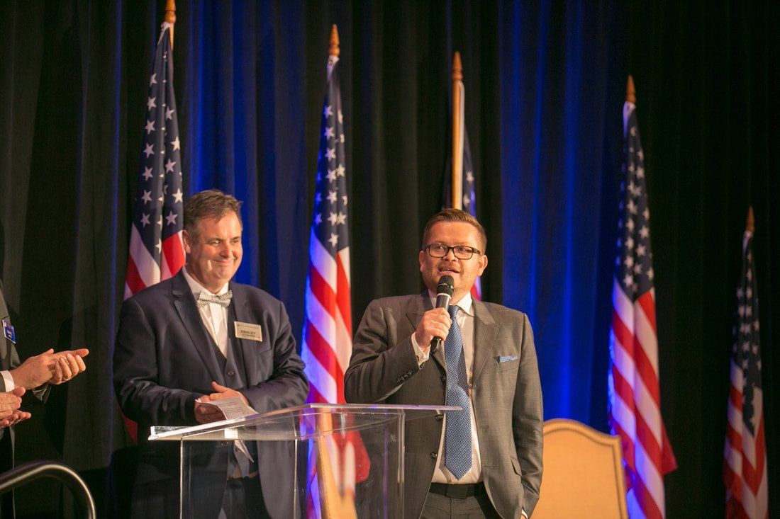 Two men are standing at a podium talking into microphones in front of american flags.