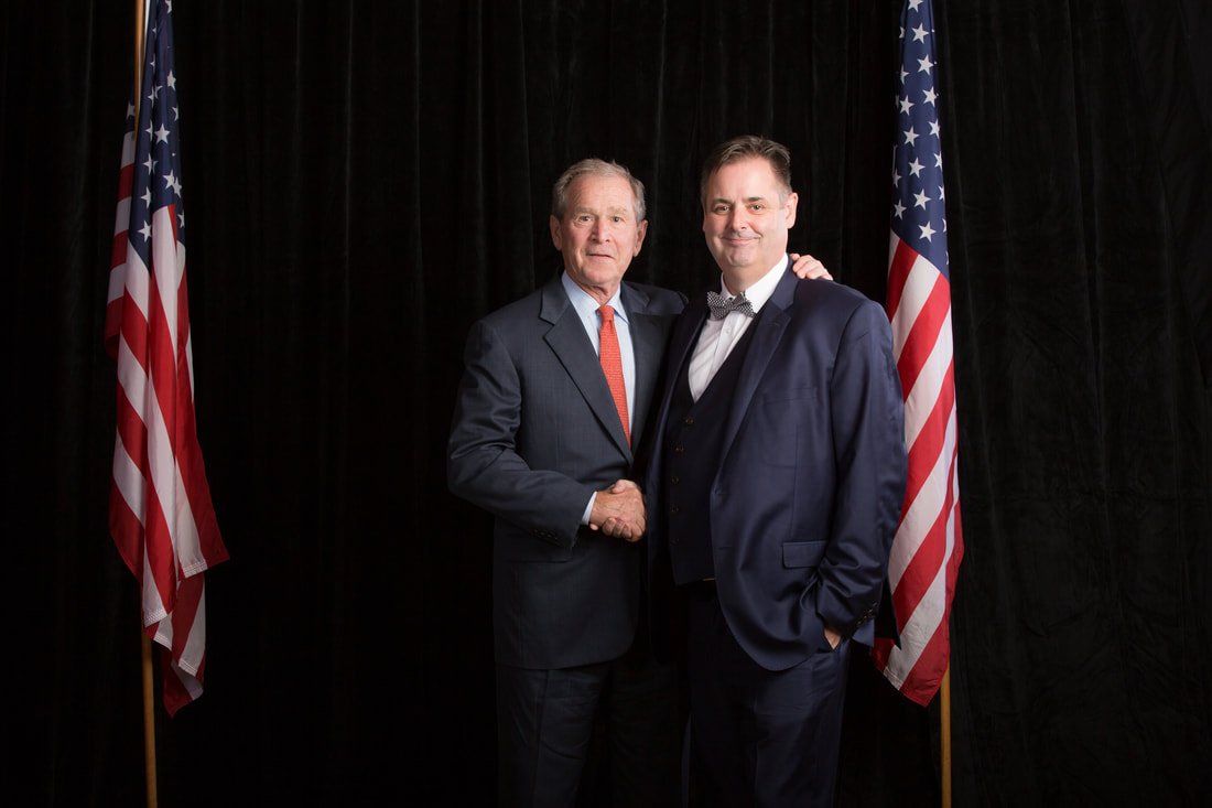 Two men in suits are shaking hands in front of two american flags.
